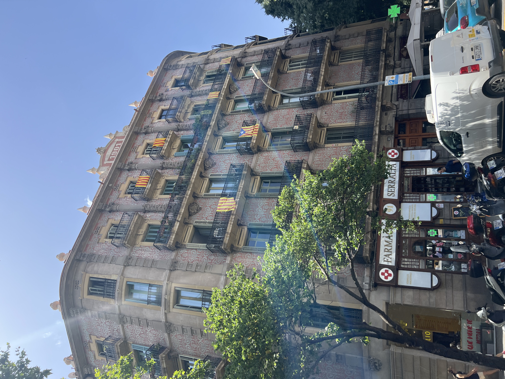This is a vertical, outdoor, eye-level medium shot of a historic building in Barcelona, Spain. The building has a distinctive architectural style with ornate detailing and decorative patterns on its facade. Several Catalan flags are visible on balconies adorned with black metal railings. Trees with lush green foliage partially obscure the lower floors of the building.
At street level, the facade transitions to storefronts. A prominent pharmacy, "FARMA SERRALIA," with a red cross symbol and the text "FORMULES MAGISTRALS" is clearly visible. A green pharmacy cross is also illuminated above the storefront. Parked on the sidewalk are several motorcycles and a white van. The white van has the license plate "4472 JWM". A blue car is also parked behind it.
The sky is clear and blue, suggesting a sunny day. The sun's rays create lens flare in the upper left portion of the image. The overall impression is one of a bright, warm day in a vibrant urban setting. The building appears to be quite old, with the year "1901" visible near the top of the facade.