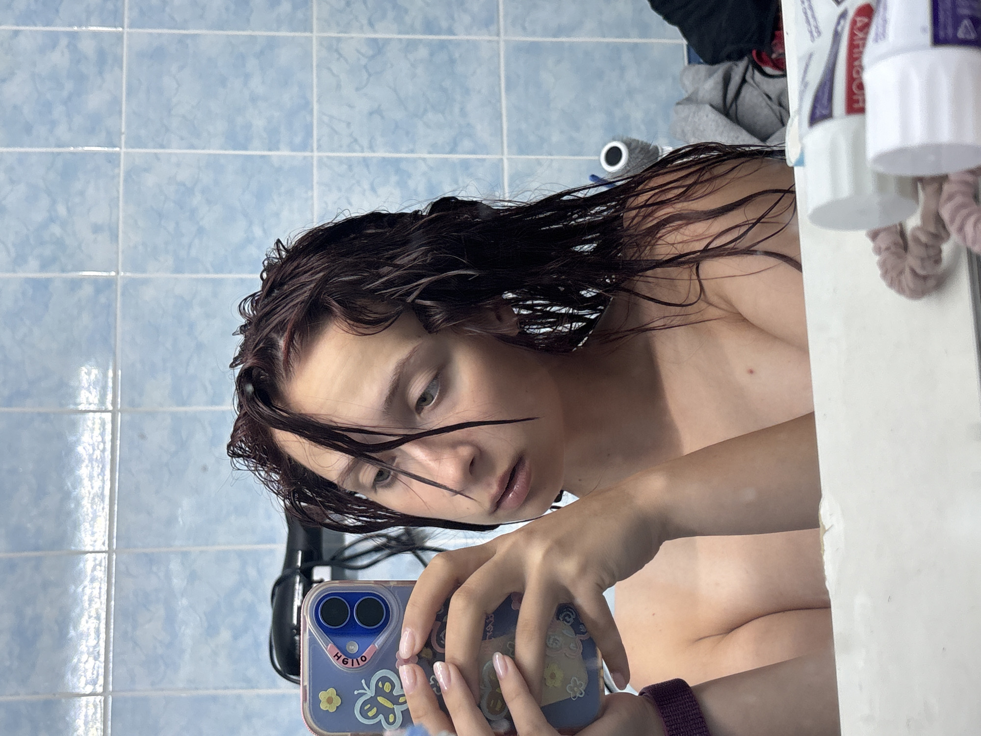 The image appears to be a self-taken photograph in a bathroom, characterized by blue tile walls. A young woman with dark, wet hair occupies the foreground, holding a smartphone in her hands to capture the image. The smartphone has a blue case adorned with stickers. The woman is looking down at the phone, partially obscuring her face. The background reveals bathroom essentials: toiletries on a shelf, and possibly clothing or towels. The location is identified as Прикамский, Russia.