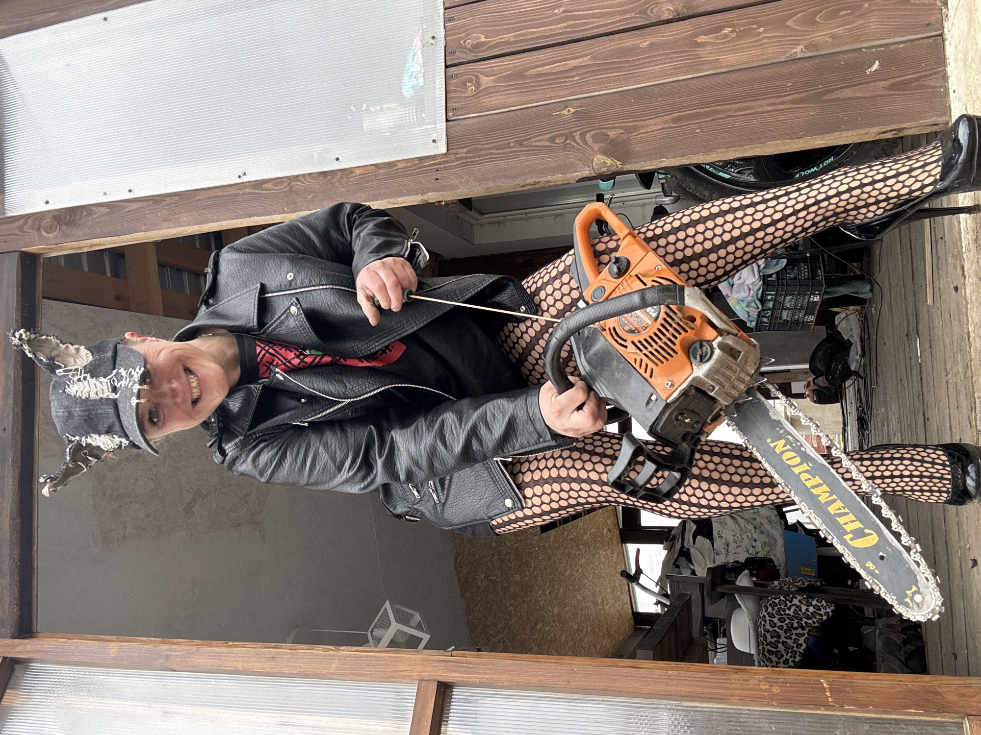 A woman, wearing a black leather jacket, a ripped denim hat, and fishnet stockings, is holding an orange and black "Champion" chainsaw. She appears to be pulling the starter cord. The location is likely a cluttered garage or enclosed patio, potentially in or near the city of Chaikovsky, Russia.