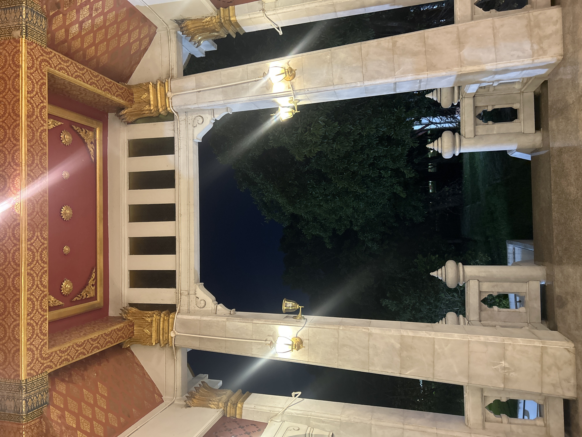 The image shows an ornate, covered entryway, likely at night, given the dark sky and artificial lighting. The ceiling is elaborately decorated with red and gold patterns, featuring intricate geometric designs and floral motifs. Large, marble-like pillars support the structure, adorned with gold accents at their capitals. Wall-mounted lights, featuring a traditional design with a glass shade, illuminate the area. Beyond the entryway, a dark expanse of foliage is visible, suggesting an outdoor environment with trees. The overall aesthetic points towards a temple or a similarly significant cultural or religious building in Thailand, potentially within Tha Khlong Town Municipality. The foreground shows a pathway or floor, also appearing to be made of stone or a similar material. There are no people visible in the image.