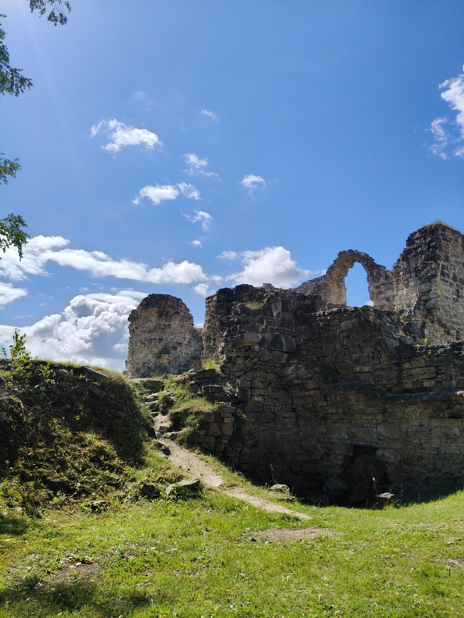 The image shows the ruins of Koknese Castle in Koknese, Latvia, on a sunny day with a blue sky and scattered white clouds. The castle ruins are constructed of stone and appear to be ancient, with some sections of walls and archways still standing. In the foreground, there is a grassy area with a dirt path leading up towards the ruins. The path winds its way through the overgrown vegetation, suggesting the passage of time and the natural reclaiming of the site. The overall impression is one of historical significance and natural beauty, with the ruins set against the backdrop of a vibrant, clear sky.