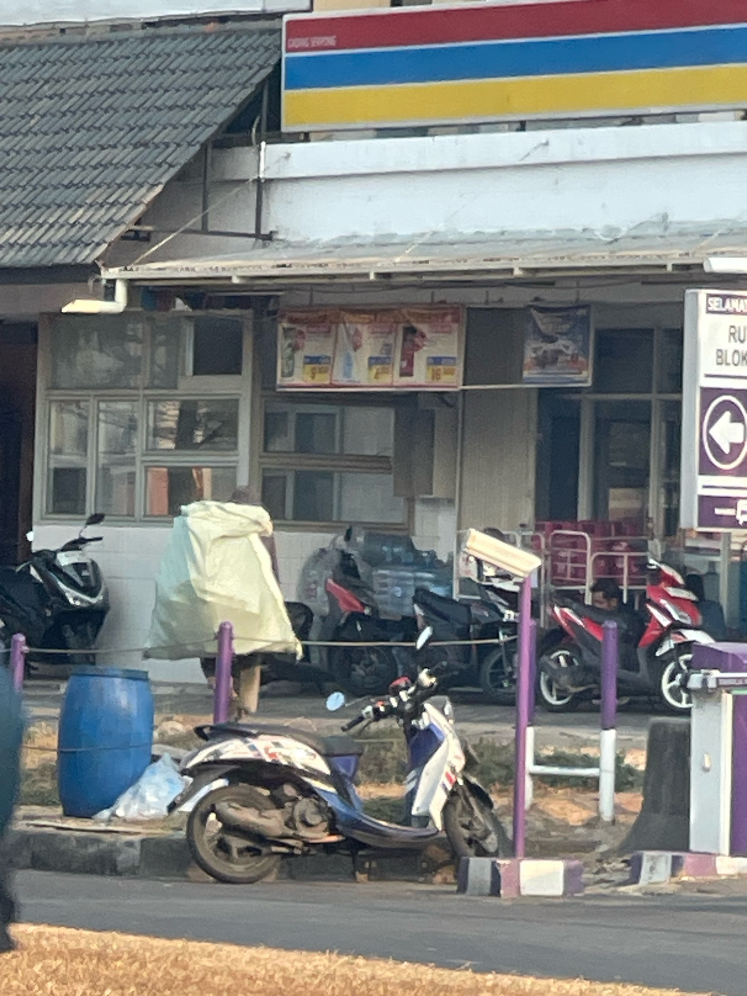 This image depicts a street scene in front of a convenience store, likely located in Pakulonan Barat, Indonesia. The store's facade features a gray tiled roof and white exterior walls. Above the entrance, a sign with horizontal red, blue, and yellow stripes is visible. Multiple motorcycles are parked in front of the store, including a black scooter on the left, a red and white scooter in the middle right, and another red scooter behind it. A person is partially obscured by a large, light-colored tarp or bag, standing near the entrance. To the left of this person, a large blue barrel sits on the ground, next to some plastic sheeting. The street in the foreground is paved and bordered by a sidewalk with purple and white striped posts. The ground beyond the sidewalk is covered in dry grass. The overall impression is a typical commercial street view in an Indonesian town, with everyday elements like parked vehicles and a convenience store.