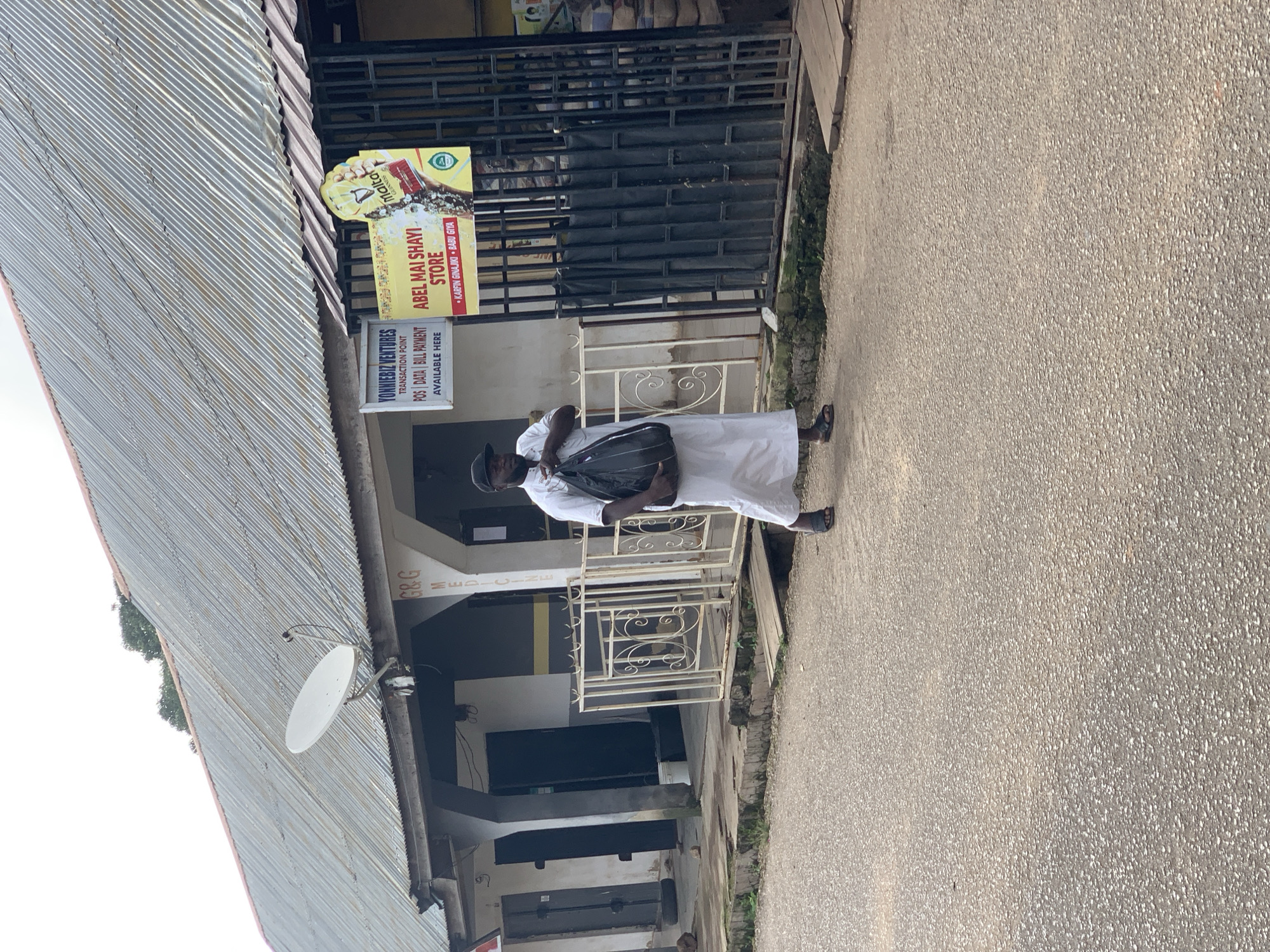 The image shows a man in a white robe and black cap walking on a paved road in front of a building in Jos, Nigeria. The building has a corrugated metal roof and several businesses are advertised on its facade, including "YONNIEBIZ VENTURES" and "ABEL MAI SHAYI STORE." A satellite dish is mounted on the roof. The man is carrying a large black bag. There is a white metal fence in front of the building, and a wooden plank extends from the sidewalk to the road. The road surface is asphalt with visible aggregate. The scene appears to be outdoors in daylight.