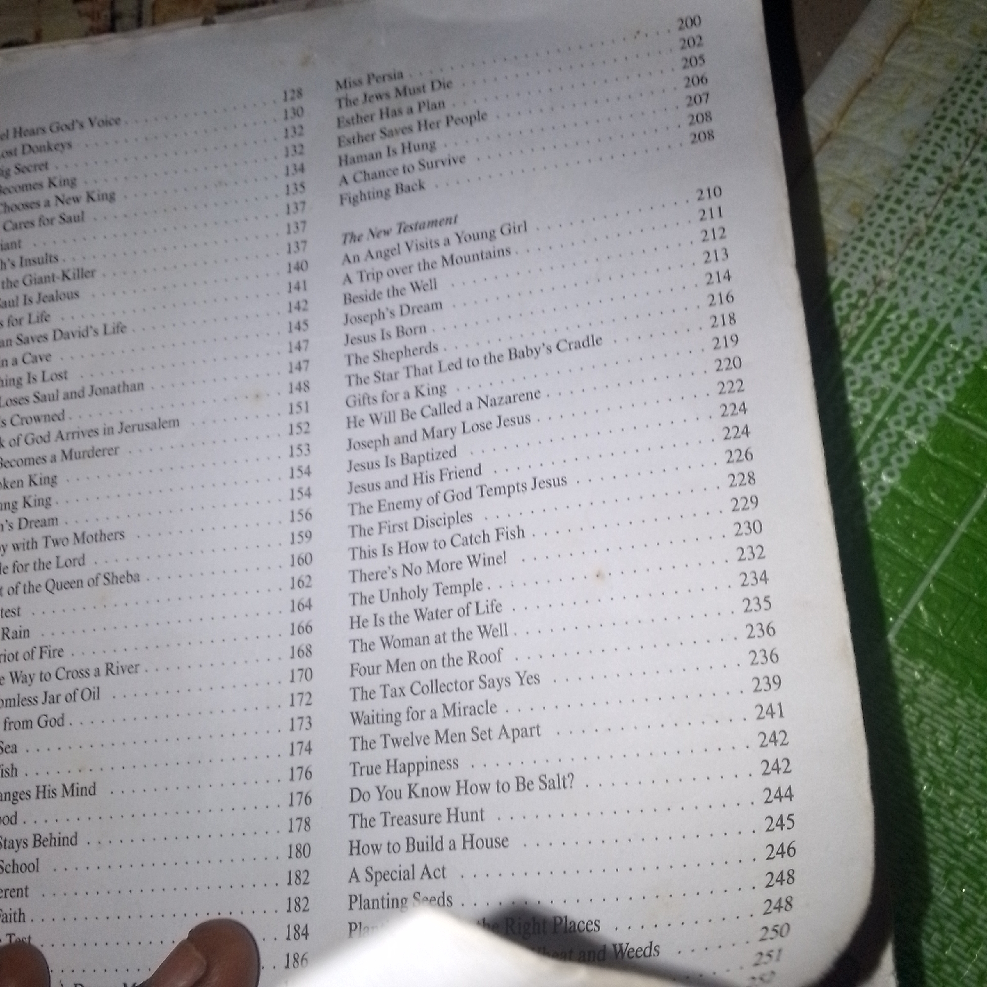 The image shows a close-up view of an open book, specifically the index or table of contents. The pages display a list of chapter titles and their corresponding page numbers, primarily in English. The text is arranged in two columns. The left column lists titles such as "Michael Hears God's Voice," "Lost Donkeys," and "Eli Becomes King," with page numbers ranging from 128 to 170. The right column begins with "Miss Persia," "The Jews Must Die," and continues with New Testament entries like "The New Testament," "An Angel Visits a Young Girl," and "Jesus Is Born," with page numbers from 200 to 251. The paper has a slightly yellowed appearance, suggesting it is an older book. The background is blurred but appears to be a textured surface, possibly fabric or a mat, with a green hue and a circular pattern. There are no people visible in the image, and the scene depicts a static object. No specific location in Jalingo, Nigeria, can be determined from the provided image.