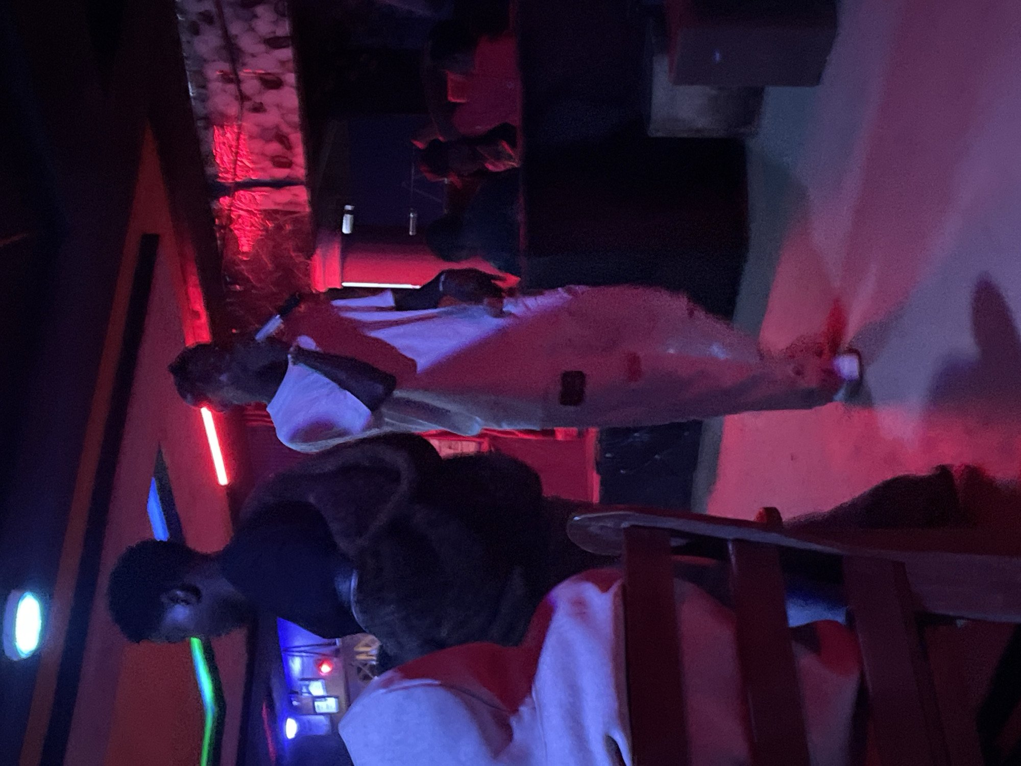 The image shows a dimly lit indoor venue, possibly a club or bar, in Durumi, Nigeria. The atmosphere is saturated with red and blue lighting, creating a moody ambiance. In the foreground, a person is seated at a wooden table with a dark surface. To the left, another individual wearing a light-colored hoodie is partially visible, also seated. In the mid-ground, a person dressed in white, holding a microphone, appears to be performing or speaking on a stage or elevated area. This individual is the central focus of the scene. Behind the performer, other patrons are visible, mostly in silhouette due to the low lighting. The environment features dark walls, possibly with decorative elements reflecting the red lights. The floor is also illuminated by the colorful lighting, casting reflections. The overall impression is of a lively night-time establishment.