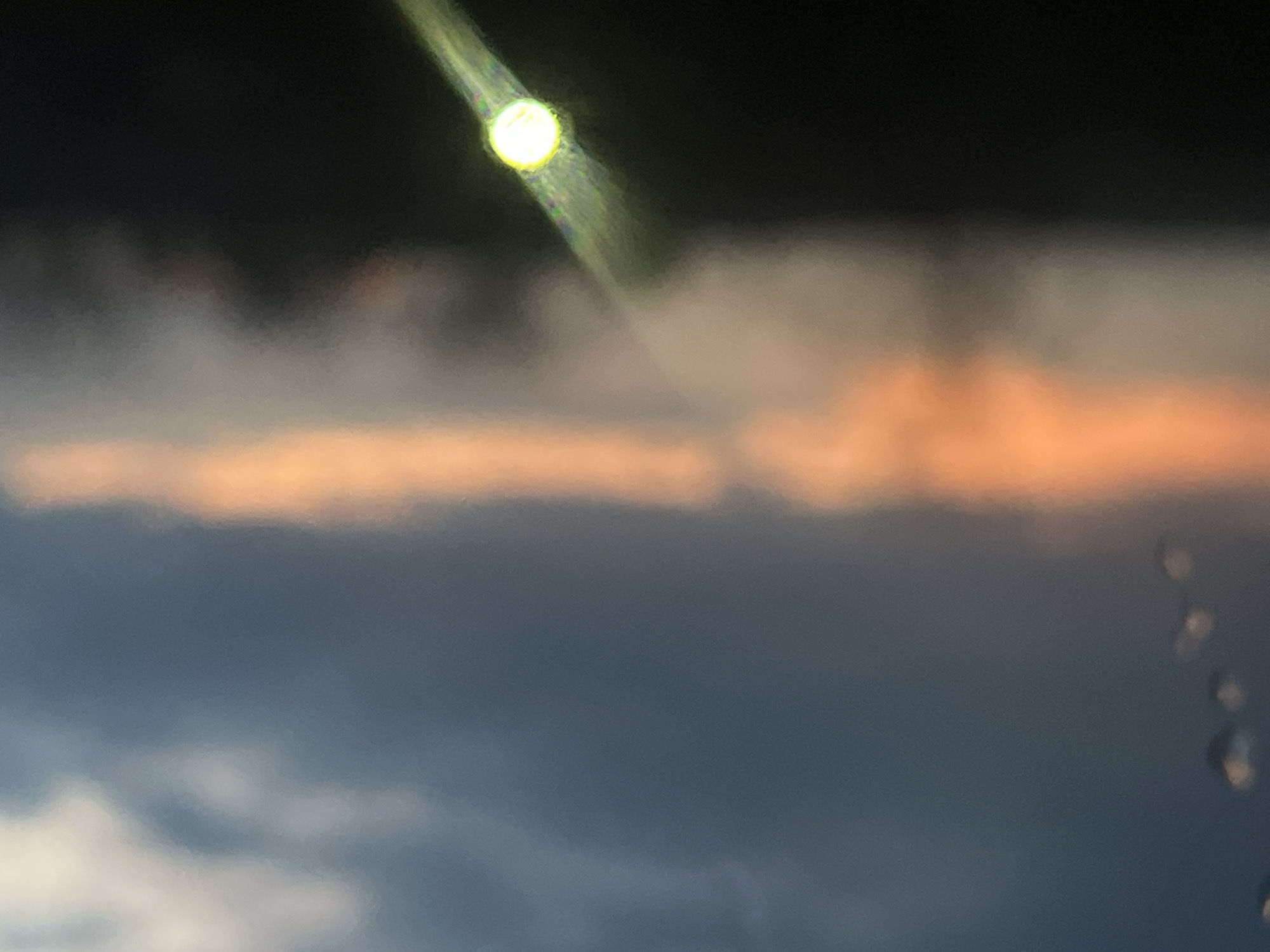 The image shows a blurred, low-angle view of a twilight sky, possibly taken through a window. The sky is dark at the top, transitioning to a band of orange-tinged clouds, and then to lower layers of blue and grey clouds. A streak of light, possibly from a street lamp or the moon, is visible at the top. There is no discernible information to relate it to the city of Votkinsk, Russia. There are no people or specific objects identified in the image.