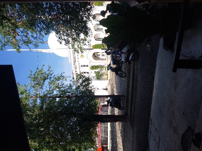 A low-angle shot reveals a white building with a dome and arched windows against a blue sky, framed by green trees on the left. A flagpole stands tall. To the right, parked motorcycles are partially obscured by shadow and foliage. A paved area leads up to the building, with a hint of a building exterior on the left and a dark structure partially obscuring the foreground on the right.