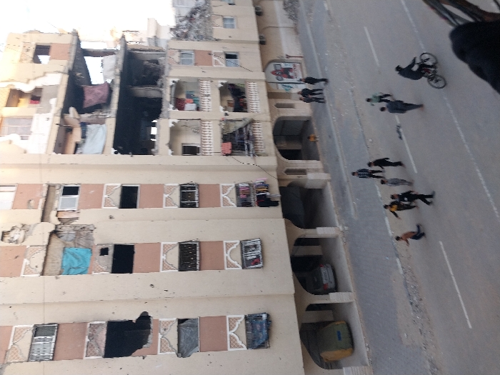 The image presents a tilted, street-level view of a severely damaged multi-story building, likely a residential apartment complex, alongside a paved road with people and vehicles.
The main subject is a large, light-colored building, appearing to have at least six to seven visible floors. It exhibits extensive structural damage: large sections of its facade, walls, and interior floors are missing or have collapsed, exposing the building's internal structure and debris. Darkened areas within the exposed sections suggest charring or fire damage. Some windows are shattered or missing, while others show hanging fabric or personal belongings, indicating recent habitation. The lower levels of the building feature recessed areas, possibly for parking, where two vehicles are visible—one dark car and another lighter, possibly yellow or beige, vehicle.
Adjacent to the damaged building is a paved road with a white dashed line marking what appears to be a lane separation. A sidewalk or shoulder separates the building from the main roadway. On the street and sidewalk, a group of approximately eight to ten people are visible. One person is riding a bicycle on the road, dressed in dark clothing. Other individuals are walking in small groups or alone; they are generally dressed in casual, dark, or neutral-colored clothing, with one person appearing to wear a yellow top. Their demeanor appears calm, suggesting they are either passing through or observing the aftermath of an event rather than being in immediate danger.
The overall scene conveys an atmosphere of destruction and aftermath. The extensive damage to the building strongly suggests a catastrophic event such as an explosion, bombing, or severe structural failure. The time of day is daytime, with natural light illuminating the scene. No discernible text is clearly visible on any signs or objects within the image.