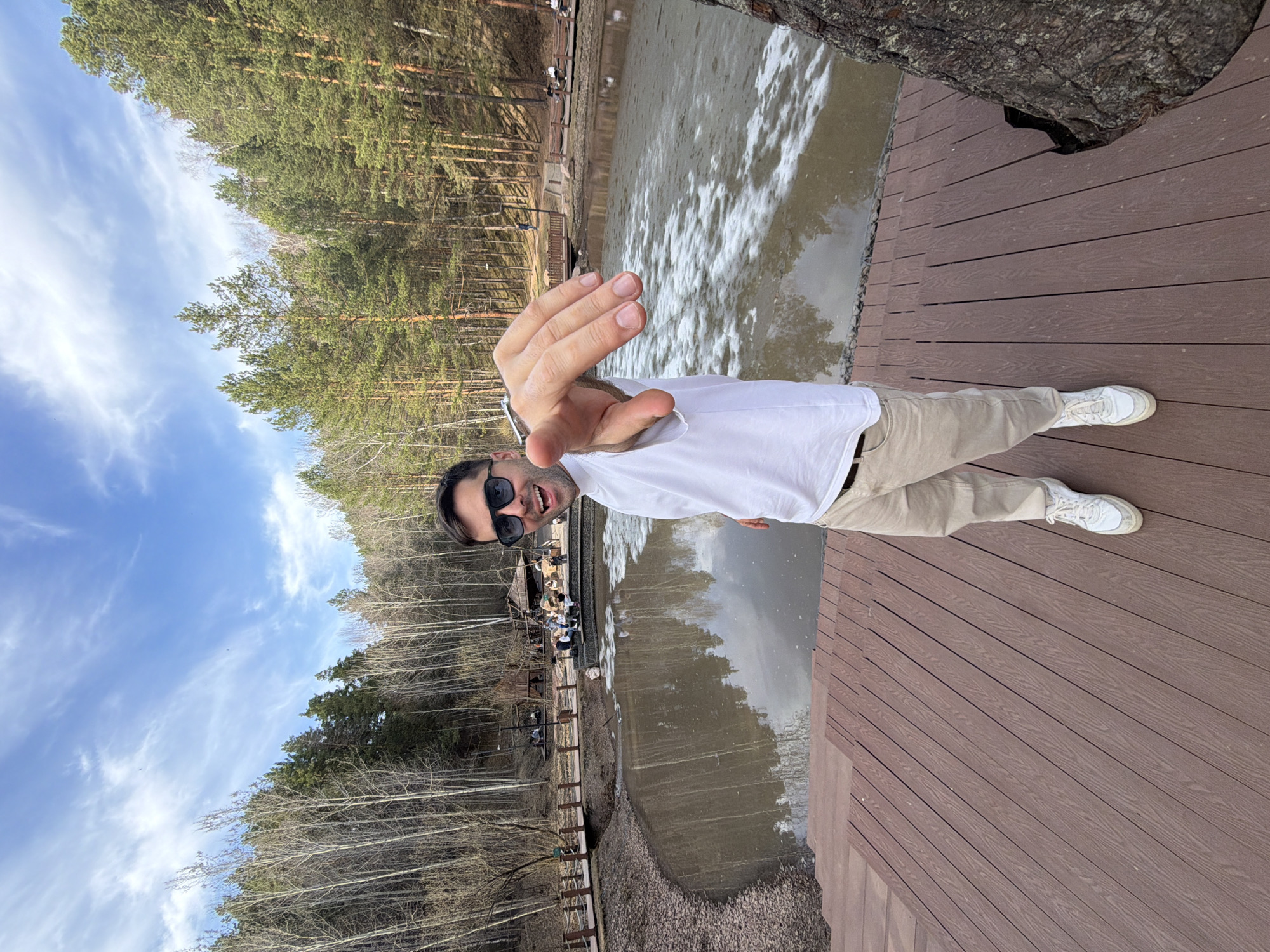 A man in sunglasses, a white t-shirt, and khaki pants extends his hand towards the camera while standing on a wooden deck. The deck overlooks a body of water, likely a pond or lake, reflecting trees on its surface. Across the water, a wooden structure houses seated people, suggesting a recreational area. The background features a forest of pine trees and a blue sky with scattered clouds. The location appears to be in a park or natural setting, potentially near Krasnoyarsk, Russia, given its characteristic Siberian landscape.