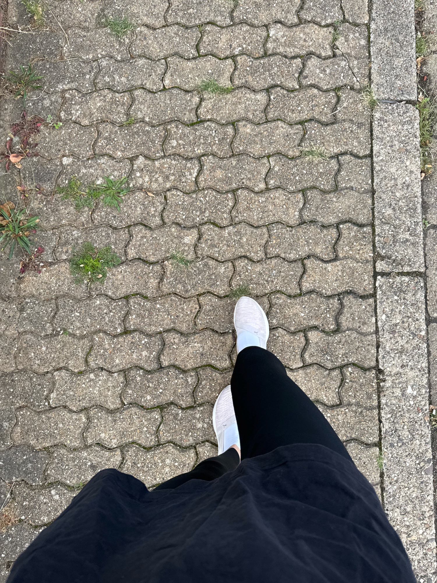 A first-person perspective shot shows a person walking on a textured, interlocking paver pathway, bordered by a concrete curb. The pathway is interspersed with patches of sparse green grass and dried leaves, suggesting an outdoor environment. The person is wearing black leggings and white athletic shoes. The scene is set in Überherrn, Germany.