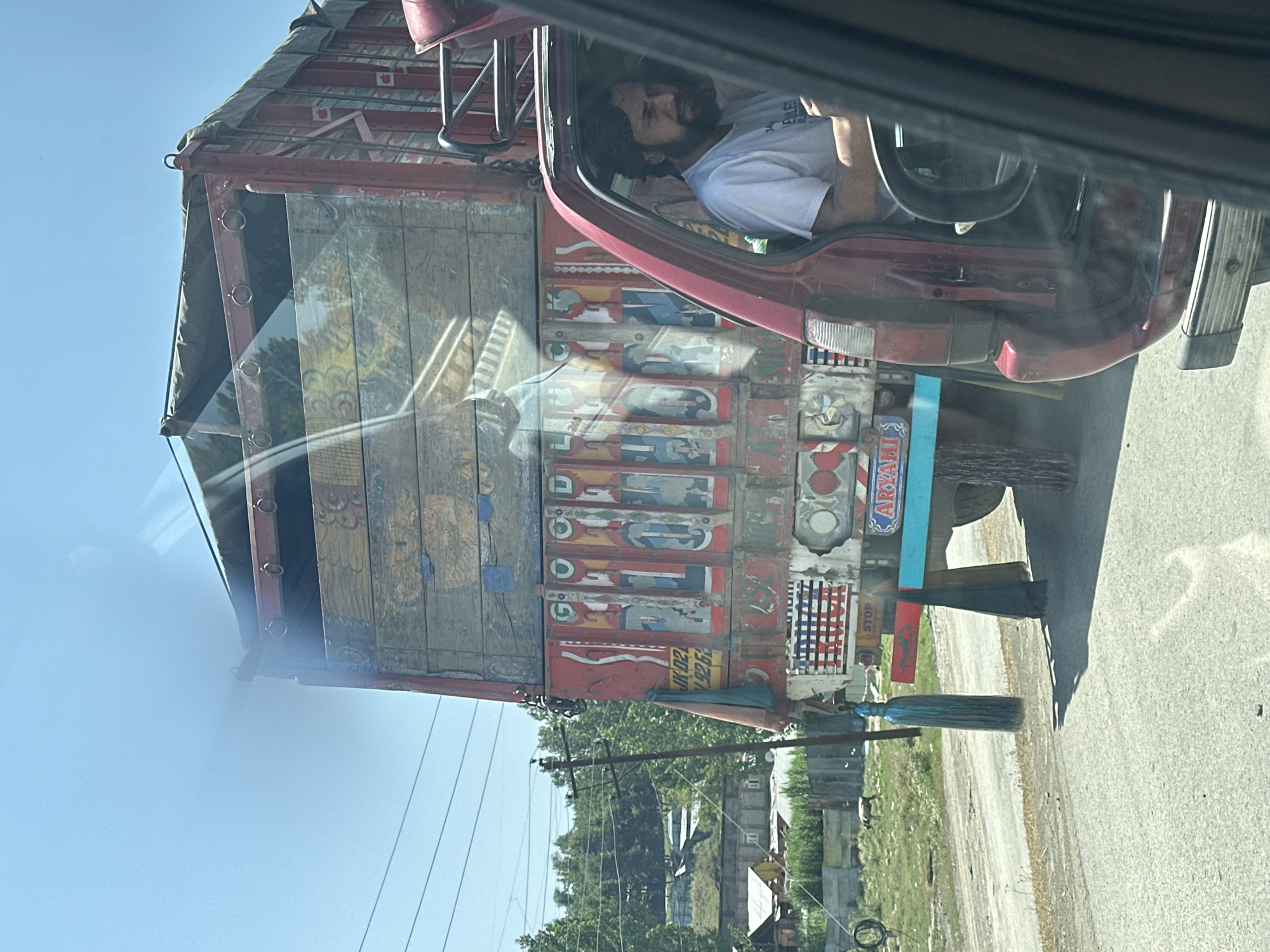 In Khrewan Lasi Pora, India, a red car is stopped on a paved road, its passenger-side door open. Inside the car, a man with a beard and wearing a white t-shirt is visible. Behind the car, a large, ornately decorated truck is parked. The truck's red and wooden body features colorful paintings and text. A teal-colored tasseled ornament hangs from the truck's rear. In the background, trees and buildings are visible under a clear blue sky. A power pole with wires stands to the left of the truck, and a few animals are in a grassy field behind a fence.