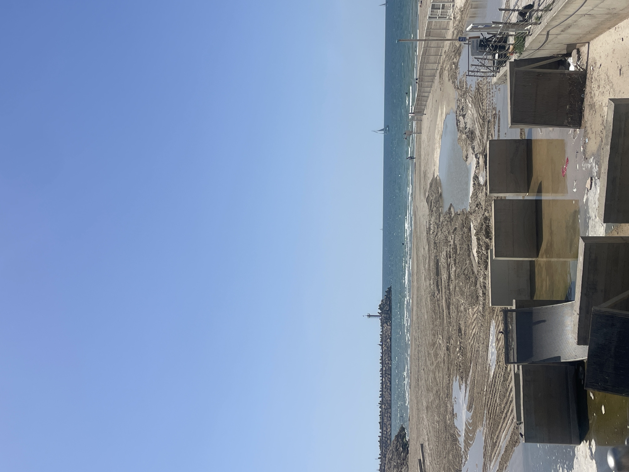 The image depicts a beach scene in Herzliya, Israel. The shoreline exhibits signs of recent heavy machinery use. Puddles of water are scattered across the sand. A breakwater extends into the sea, and a lighthouse is visible. In the foreground, the top of a set of concrete steps is partially visible. The sky is clear and blue.