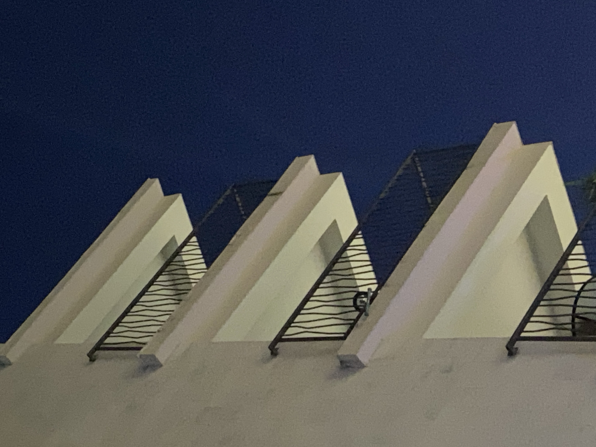 A low-angle shot shows three white, triangular architectural structures against a dark blue sky, likely at night. The structures appear to be part of a building's facade, with dark, wavy metal railings or grilles filling the triangular spaces within. The building's surface is a muted white color. The photograph does not include any people, specific actions, or dynamic interactions, and its location is not identifiable from this image alone in terms of the City of Budva, Montenegro.