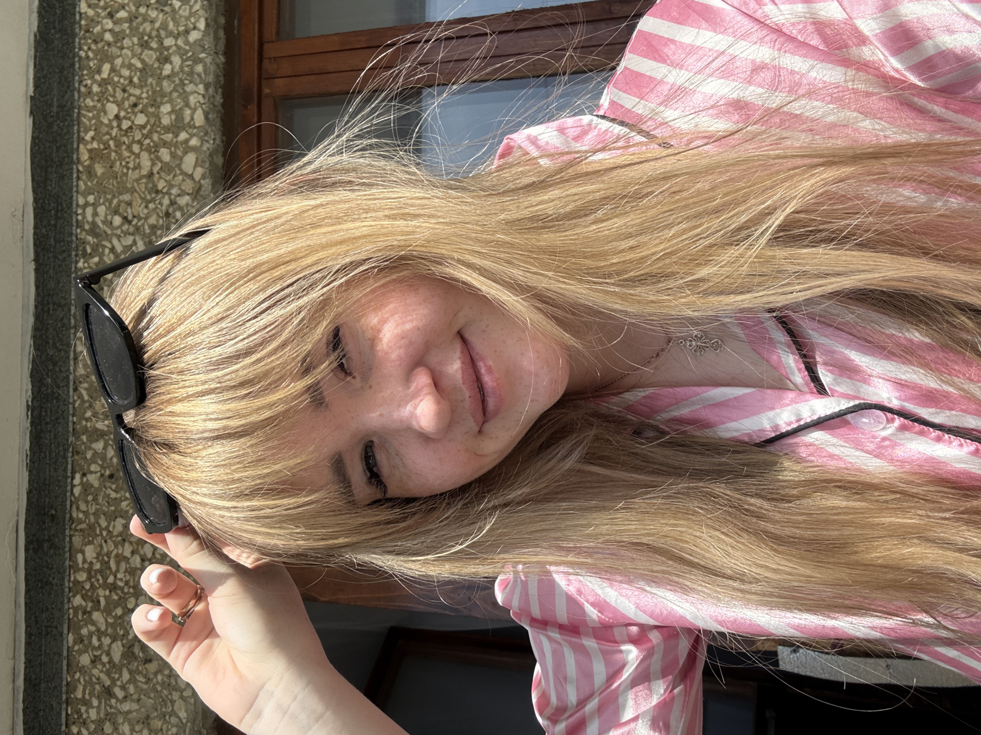 A young woman with long, light blonde hair is posing for a photo. She is wearing a pink and white striped pajama top with black piping. Sunglasses are perched on top of her head. She is wearing a ring on her left ring finger. The background appears to be an exterior wall and a window. The image was taken in the city of Марковский, Russia.