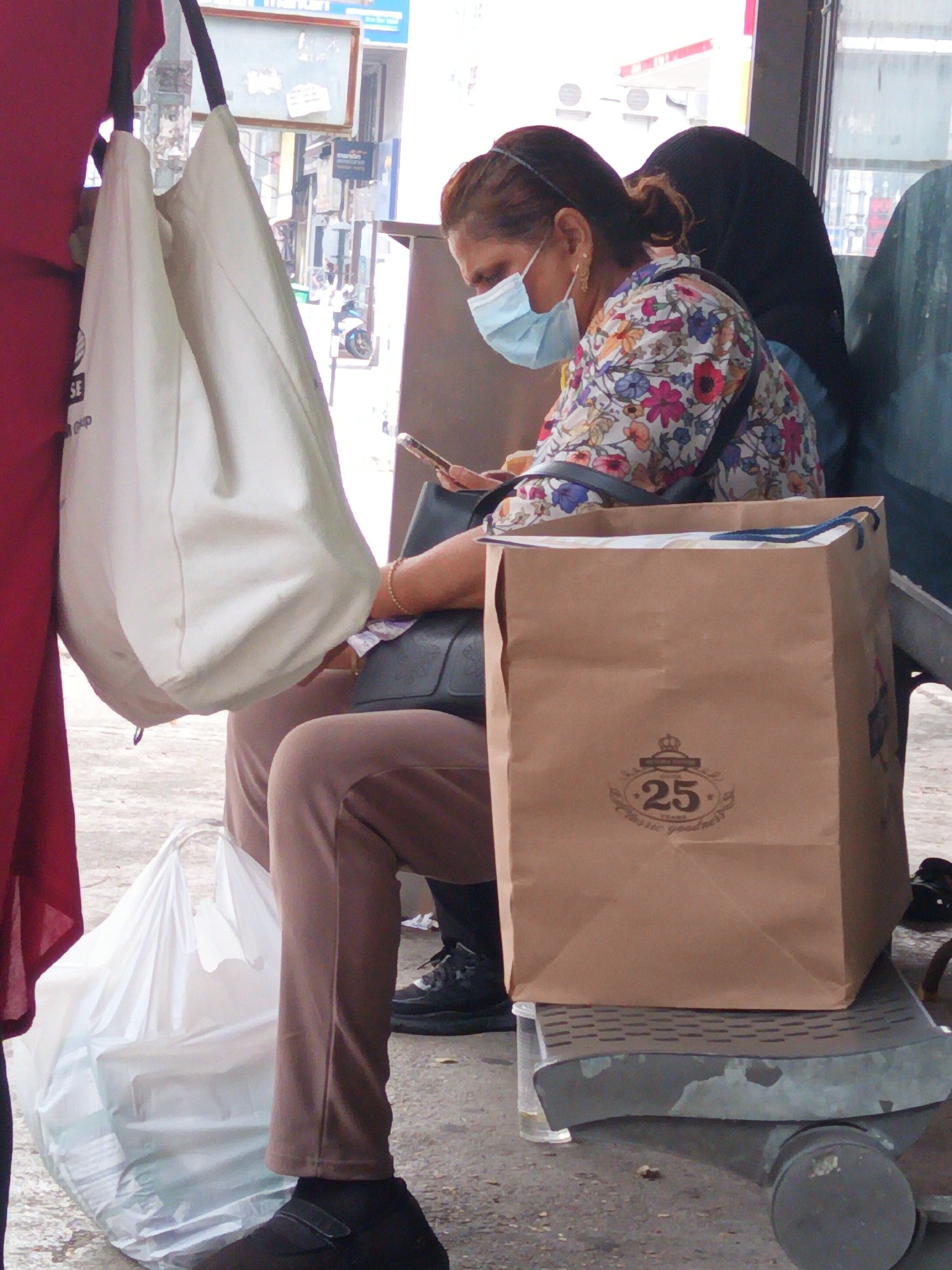 The image depicts a woman wearing a floral-patterned shirt and a light blue face mask, sitting on a bench outdoors. She is holding a mobile phone in her right hand and has a dark-colored bag on her lap. Beside her, a large, light-colored fabric tote bag hangs from a hook on a red-colored structure. In front of her, a brown paper shopping bag with a "25 YEARS Classic goodness" logo rests on the bench. A white plastic shopping bag is visible on the ground to her left. The background shows a street scene with buildings and a motorcycle, suggesting an urban environment. The presence of a bench, shopping bags, and people in a public area indicates a bus stop or waiting area in a city. The overall scene suggests a moment of waiting or transit in a public space in Kuala Lumpur, Malaysia.