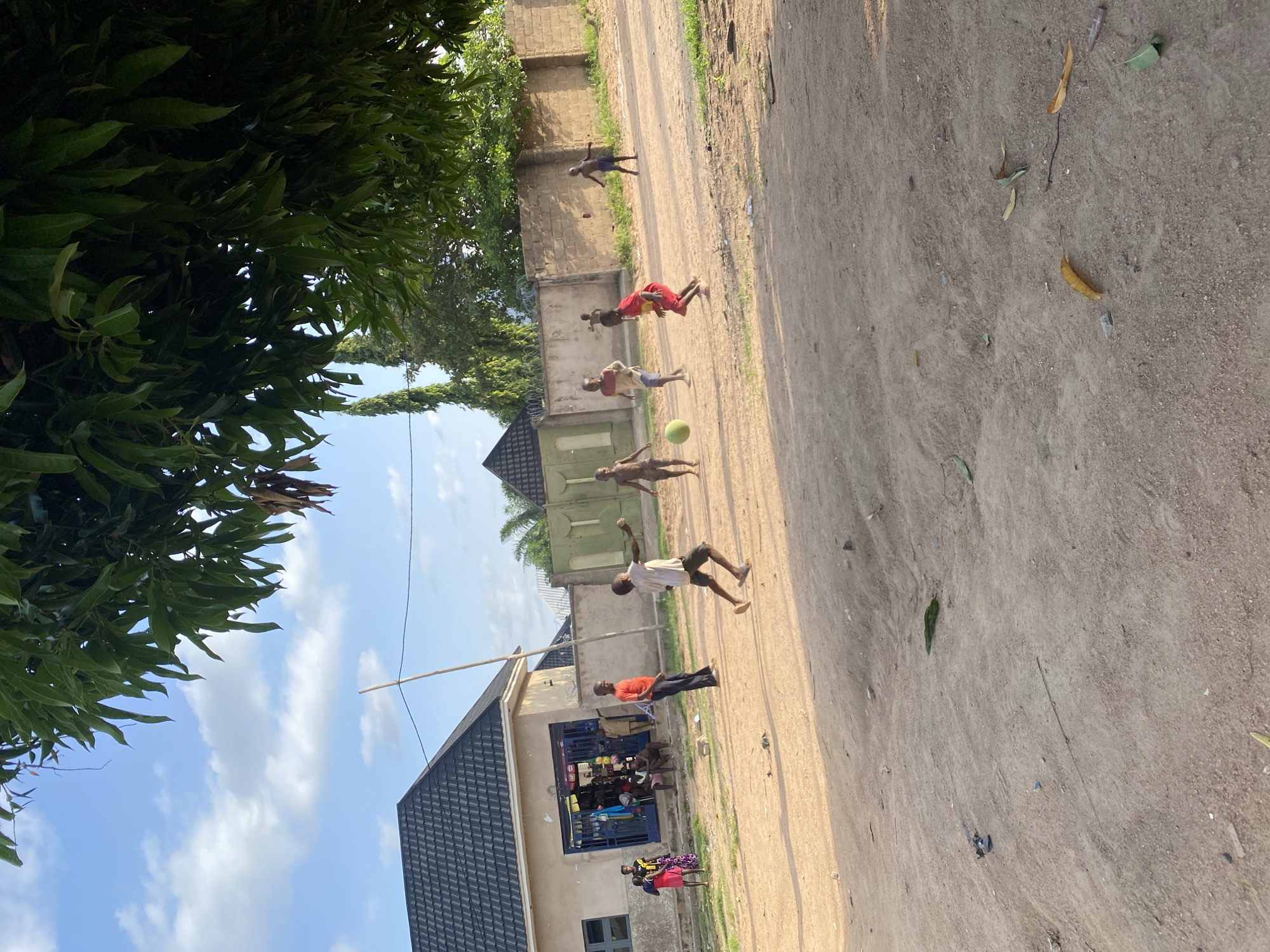 This image depicts a lively scene in Howayi, Nigeria, likely on a sunny afternoon given the bright lighting and clear sky. The primary subjects are a group of children actively playing soccer in a dirt road. Several children are in motion, one mid-kick with the ball, others running or poised to play. The setting is a somewhat dusty, unpaved road lined with buildings and vegetation.
To the left of the road, lush green trees partially obscure the view, with their leaves creating a dappled light effect on the ground. Behind the trees, a brick wall and some fencing are visible. On the right side of the road, there are several structures. One building appears to be a small shop with its blue-painted entrance open, displaying various items. Other buildings with dark tiled roofs and neutral-colored walls are also present.
The children are dressed in casual, colorful clothing, a mix of t-shirts and shorts or dresses. Their energetic poses suggest a joyful and engaged atmosphere. The time of day appears to be afternoon, with the sun casting shadows and illuminating the scene. There is no visible text in the image. The overall impression is one of everyday life and childhood play in a vibrant community.