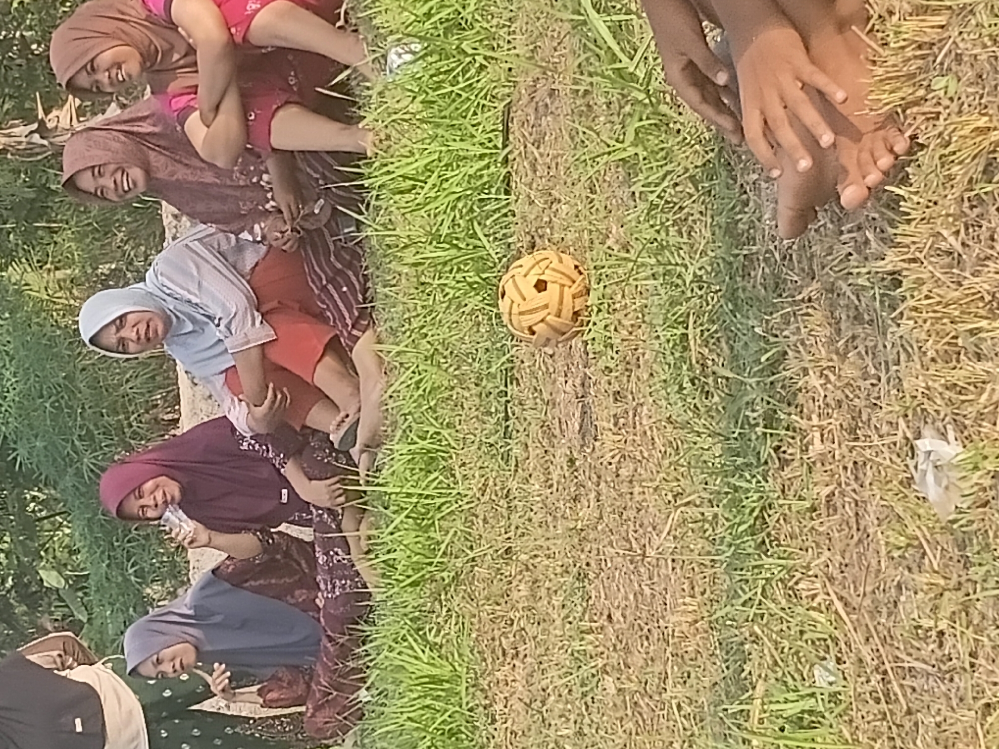In Margo Rukun, Indonesia, a group of five women are seated on the ground in a grassy area. Four of the women are visible from the waist up, and one woman is visible from the knees down. The women are all wearing hijabs and appear to be observing an event. One woman is drinking from a clear plastic cup, another is laughing, and the other three are looking towards the side. A rattan ball, used in the sport of sepak takraw, is placed on the ground in front of them. The background consists of green foliage and a dirt path. The lighting suggests it is daytime, likely late afternoon, given the long shadows.