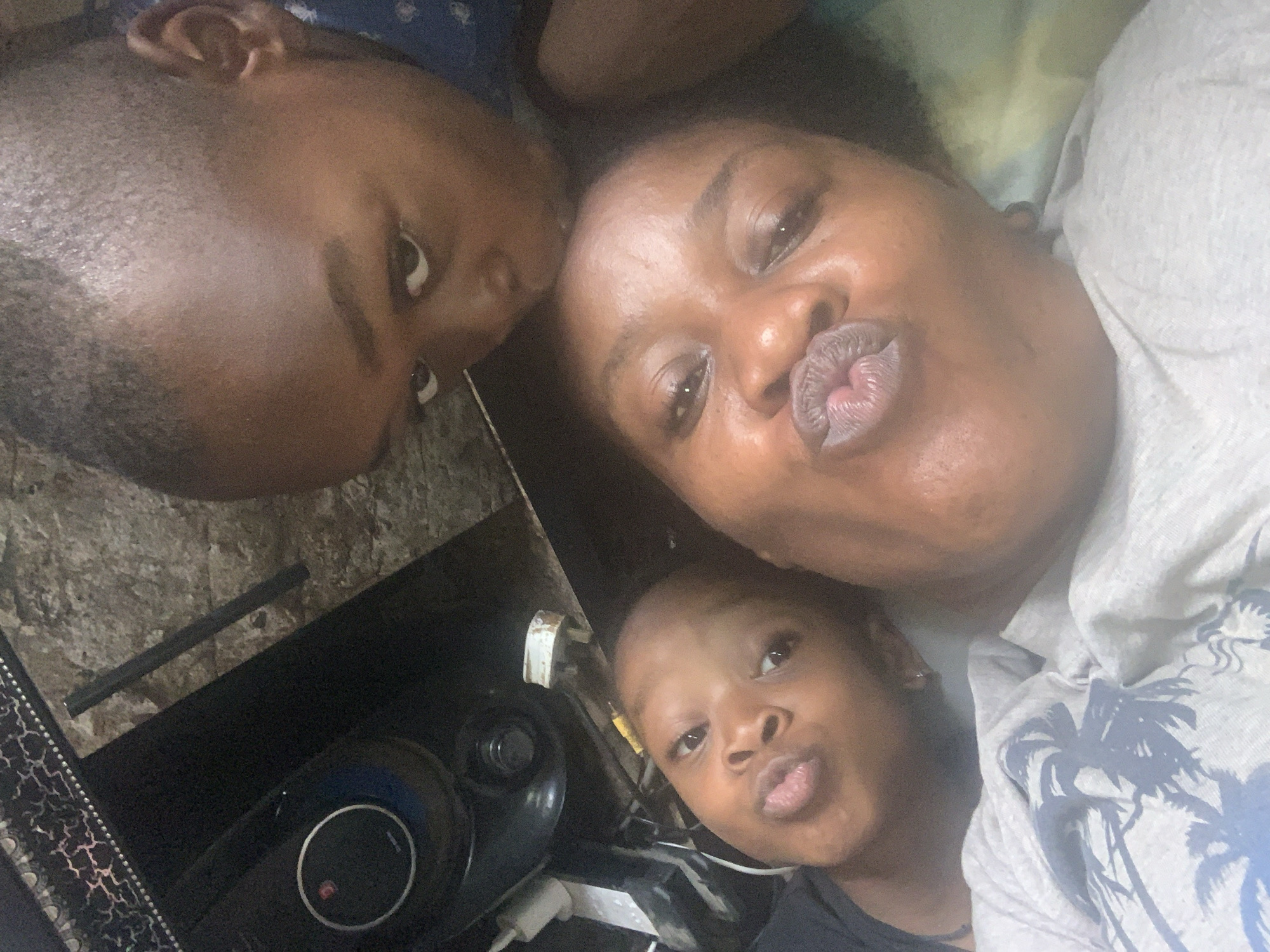 Three individuals appear to be positioned closely together, potentially for a selfie. An adult female with dark hair is wearing a light-colored t-shirt with a palm tree design. She is making a kissing face. To her left, a young child with short hair is also making a kissing face. Above the woman is another child with short hair, looking toward the camera. A black object is visible with a white cable plugged into it. The background is indistinct. There is no verifiable information indicating this photo was taken in Sabon Tasha, Nigeria.