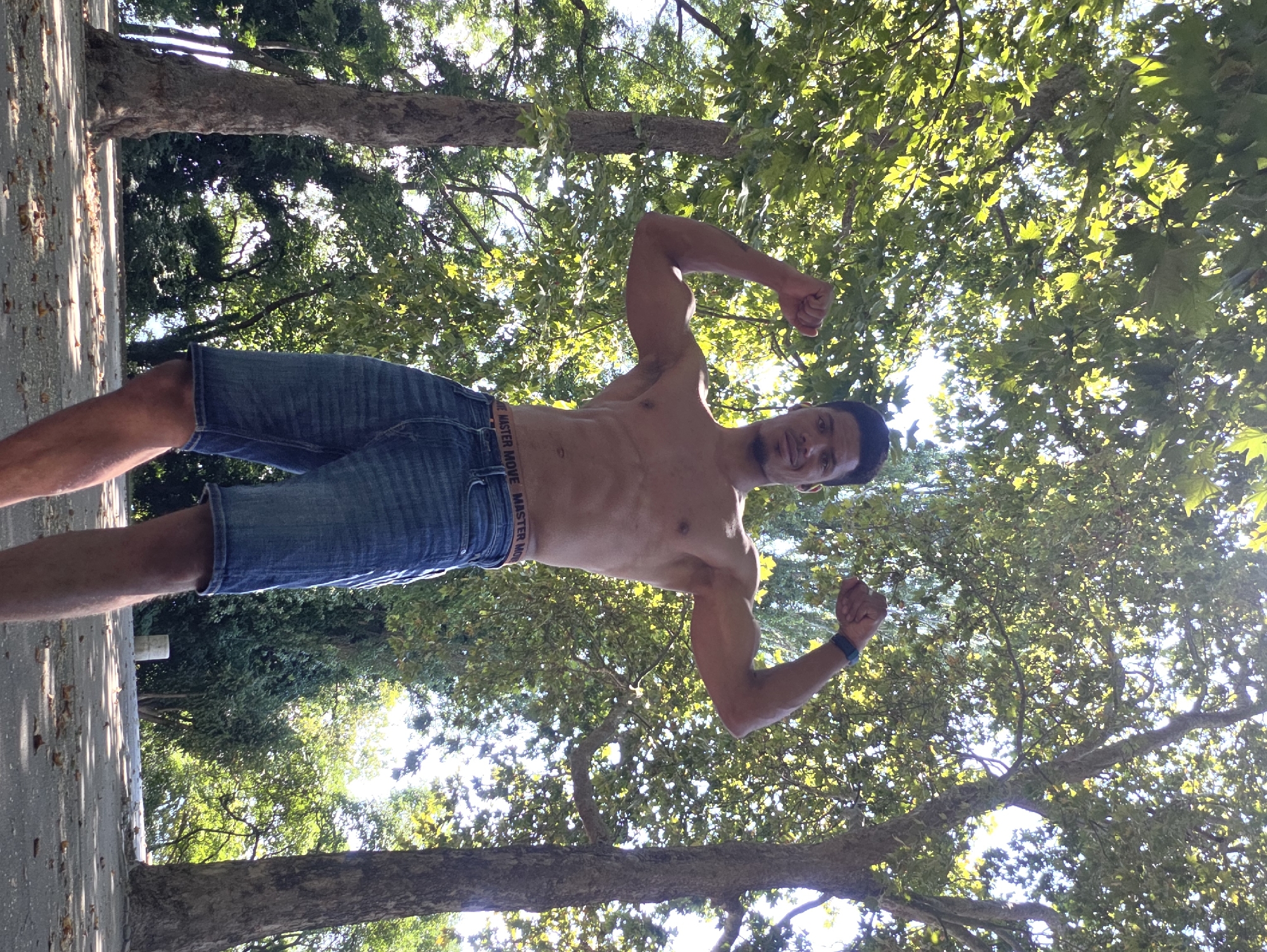 A man with dark hair and brown skin is standing outdoors, posing shirtless with his arms flexed. He is wearing denim shorts and a watch on his left wrist. The background is filled with lush green trees and sunlight filtering through the leaves. The ground is paved and scattered with fallen leaves, suggesting an autumn or late summer setting. The scene appears to be in a park or a wooded area within the city of Varna, Bulgaria.