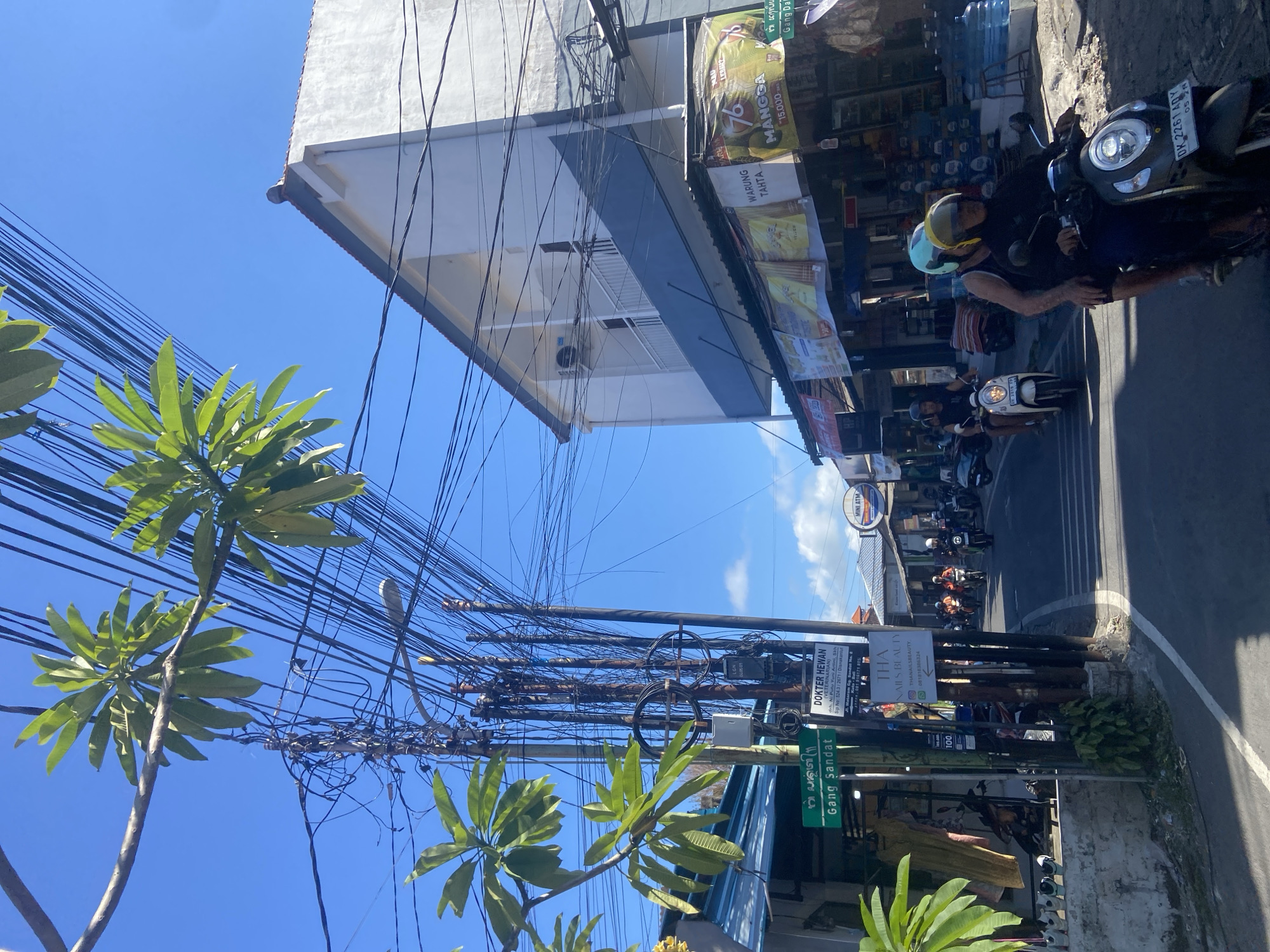 The image depicts a street scene in Kuta, Indonesia. In the lower left, the street sign reads "Gang Sandat," indicating the specific location. A complex network of overhead power lines dominates the view above, intertwined with the fronds of plumeria trees. The right side of the image shows a line of parked scooters along the curb, with a few riders visible. Several businesses line the street, identifiable by signs and storefront displays, including "Warung Tahta" and "Tha Nails Beauty." Additional signage mentions a veterinary clinic, "Dokter Hewan."