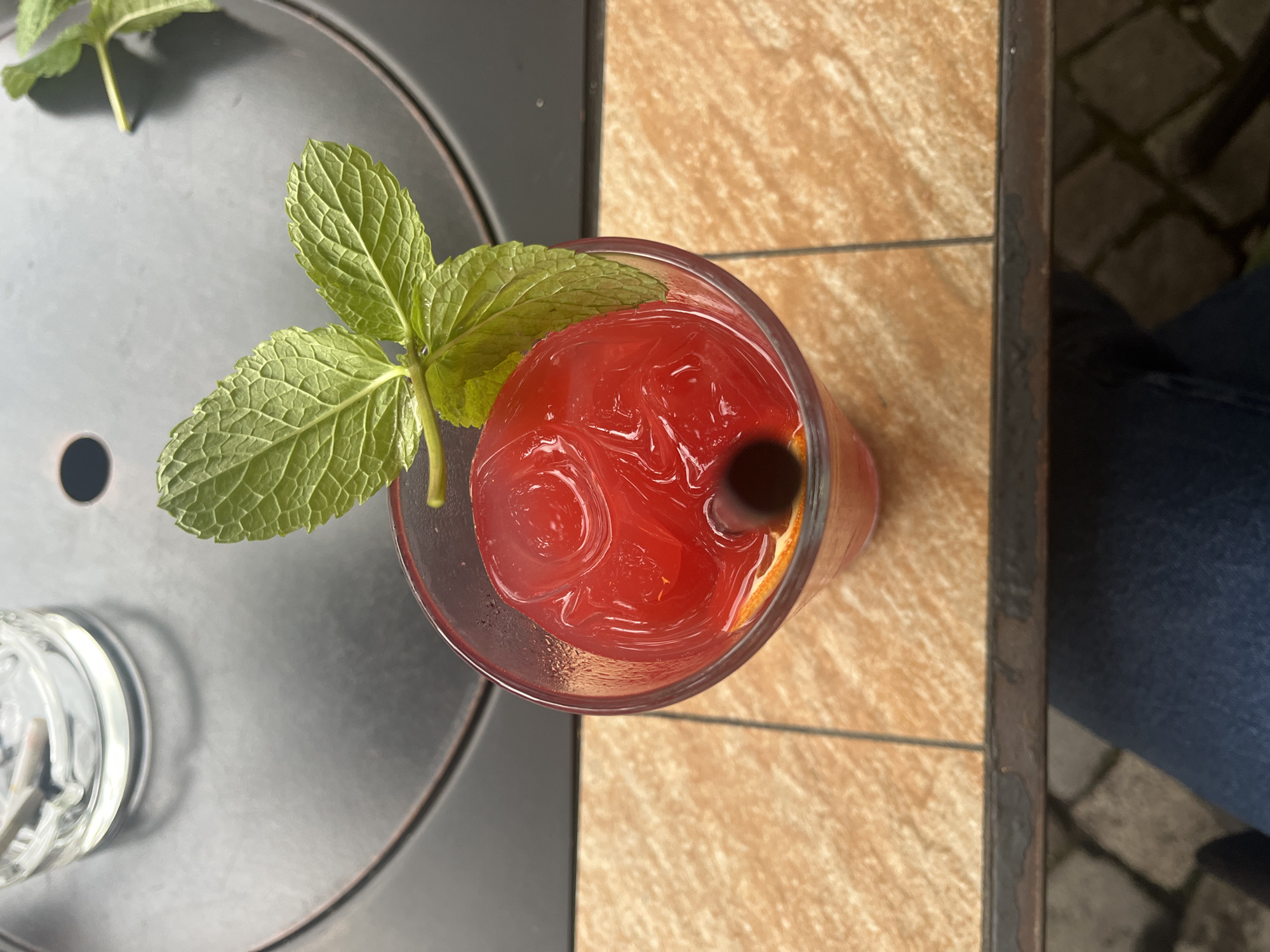 An overhead shot captures a red cocktail in a glass, garnished with a sprig of mint and containing ice, sitting on a table. A dark straw protrudes from the drink. The table surface is split between a dark grey, circular surface and a tan tiled area. A leg and pants are visible on the right side of the image, likely belonging to a person sitting at the table. The setting is possibly outdoors, given the natural light and the glimpse of stone paving to the right. Without additional context, pinpointing the exact location within Plovdiv, Bulgaria is not possible.