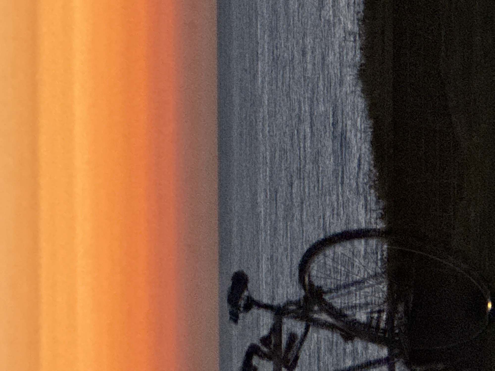 The image captures a serene coastal scene at dusk or dawn. The sky exhibits a gradient of warm orange and yellow hues, suggesting the setting or rising sun. Below the sky, a calm, grayish body of water, likely the sea, stretches across the frame, with subtle horizontal lines indicating gentle ripples on its surface. In the foreground, a bicycle is silhouetted against the background. The bicycle is dark in color and appears to be a road bike or hybrid, with its frame, handlebars, seat, and wheels visible. It is positioned on a grassy or vegetated area that slopes downwards towards the water. The overall impression is one of tranquility and natural beauty. Given the context of Tel Aviv, Israel, this scene likely depicts a location along its Mediterranean coastline, such as Jaffa Port or a beach promenade, where such views are common. There are no people or animals visible in the image.