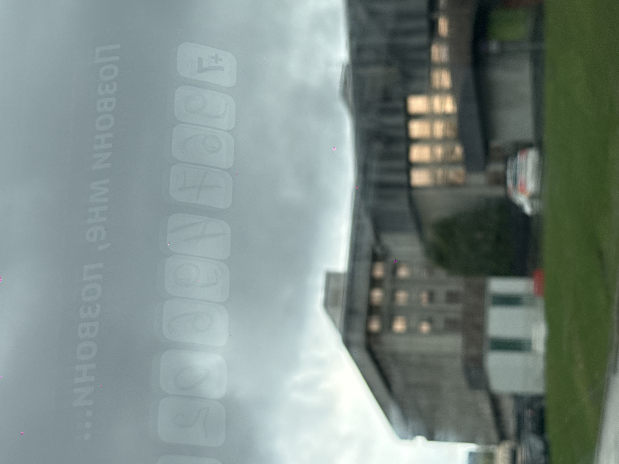 The image shows a view through a window, likely from a vehicle, with a reflection of the vehicle's infotainment screen. Reflected on the screen are various app icons. The external view is of a cityscape, including a building with visible windows and a green area, possibly a park or field. The city is identified as Moscow, Russia.
