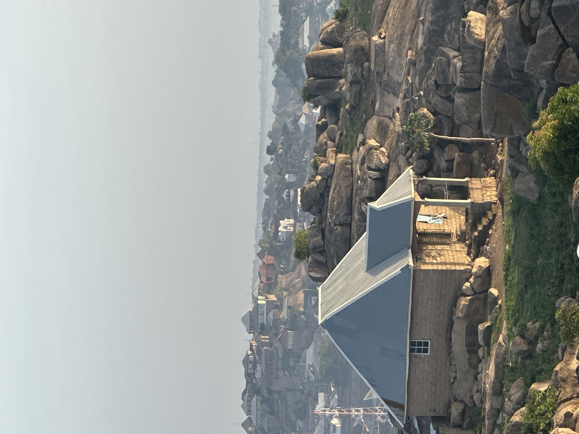 This high-angle shot, likely taken from a higher vantage point in Jos, Nigeria, showcases a unique blend of natural rocky landscape and human settlement. In the mid-ground, a stone or concrete building with a prominent light gray gabled roof stands amidst large, weathered rock formations. This house features a small, rectangular window and a balcony or terrace, where a small, light-colored figure, possibly a statue, is vaguely visible.
The scene is characterized by numerous irregular rock outcrops, some adorned with sparse green vegetation, suggesting a rugged, perhaps semi-arid environment. Behind the main house, a simple wooden bridge or walkway can be seen spanning between two rock formations. A metal railing is also visible on a paved terrace adjacent to the house.
The background extends into a sprawling urban or semi-urban area, with countless buildings stretching across the terrain. This distant townscape is partially obscured by a soft, atmospheric haze, contributing to a muted palette. The bright, hazy sky indicates daytime, likely late morning or early afternoon. The overall impression is of a community closely integrated with its rocky natural surroundings. No specific activities are clearly visible, and there is no discernible text in the image.