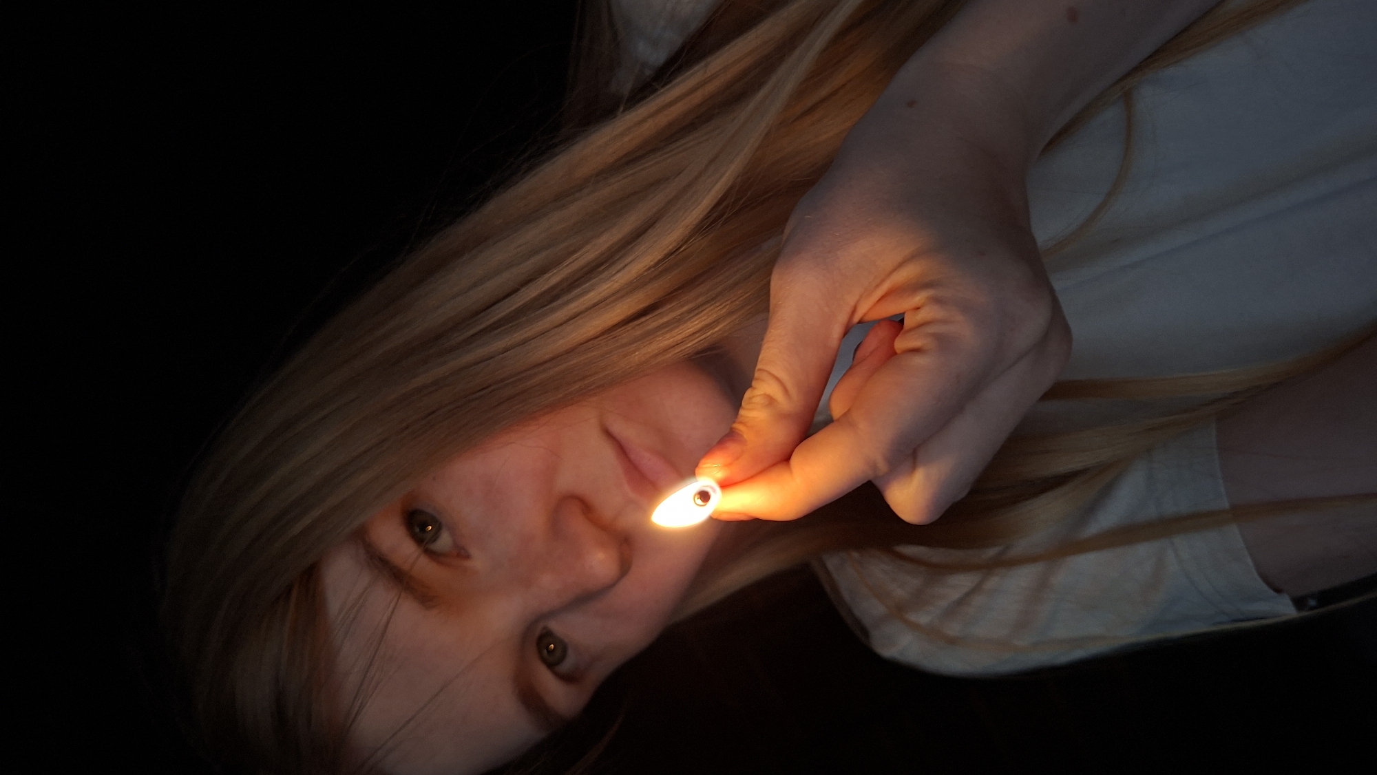 A young woman with long blonde hair is holding a lit match in her right hand. The match is positioned in front of her face, with the flame illuminating her features and casting a warm glow on her hand and neck. She is wearing a light-colored shirt. The background is dark and indistinct. The image appears to have been taken indoors in low light conditions. There is no information available about the location in the city of Большой Букор, Russia.