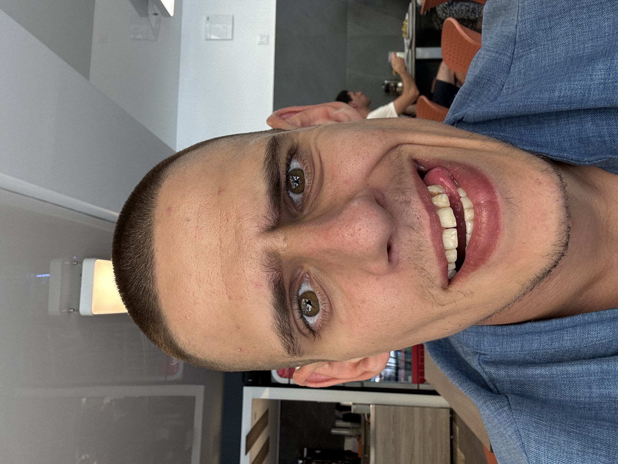 A man with a shaved head and a denim shirt is taking a selfie in what appears to be a cafe in Sofia, Bulgaria. His expression is playful as he sticks his tongue out, revealing his teeth. In the background, other patrons are visible, some eating at tables. The cafe has a modern design with hanging lights and dark gray walls.