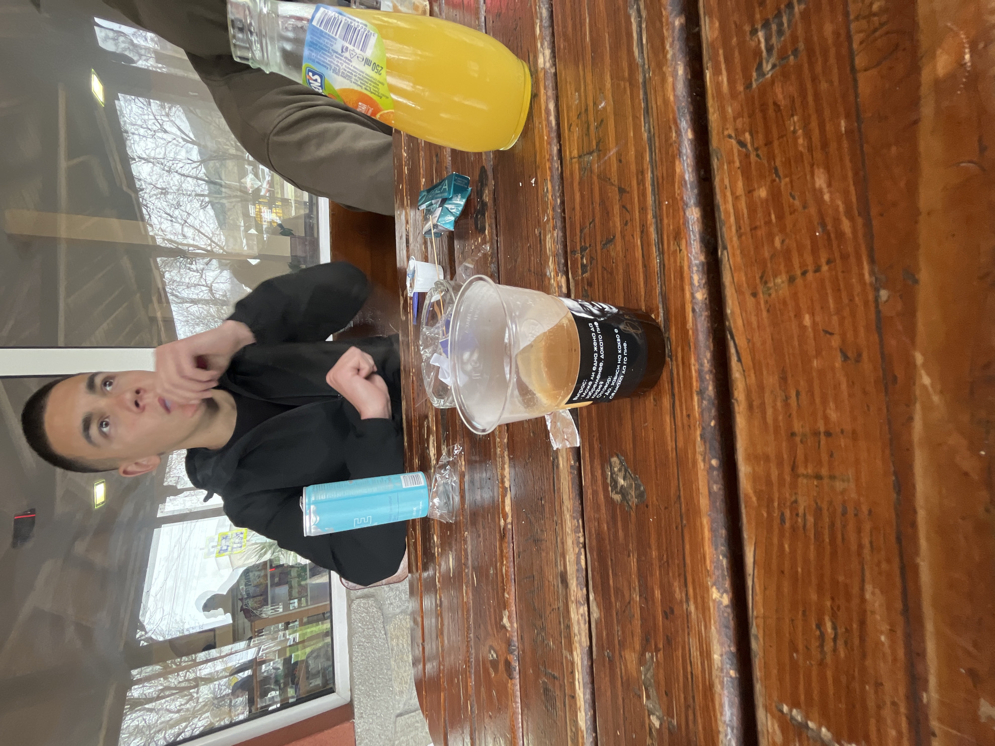 The image depicts a scene at a cafe or restaurant in Asenovgrad, Bulgaria. A young man with short dark hair is seated, wearing a black hoodie. On the wooden table, there's a glass of coffee, a can, a bottle of juice, and some discarded packaging. The background shows a window with a reflection of the outside scene, including trees and buildings. The OCR text indicates Bulgarian text.