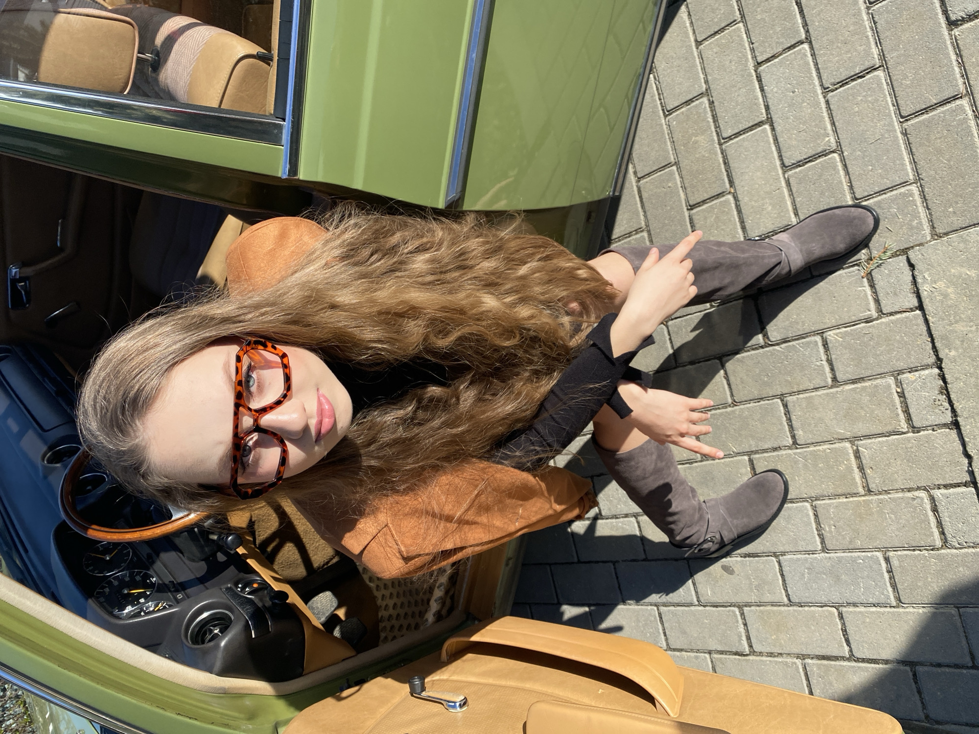 A young woman is positioned within the driver's side door of a green vintage car, likely a Mercedes-Benz based on the interior. She wears distinctive tortoise-shell glasses, a black top, and a tan jacket, and gray suede knee-high boots. Her long brown hair is loose. She is sitting in the doorway of the car with her legs extended onto a paved area made of gray rectangular bricks. The car's interior features tan upholstery and a wood-rimmed steering wheel. The location may be in a public area in Yekaterinburg, Russia, though there are no distinct landmarks visible to confirm this. The scene is well-lit, suggesting an outdoor environment. The angle of the photograph is a high-angle shot, looking down at the subject and the scene.