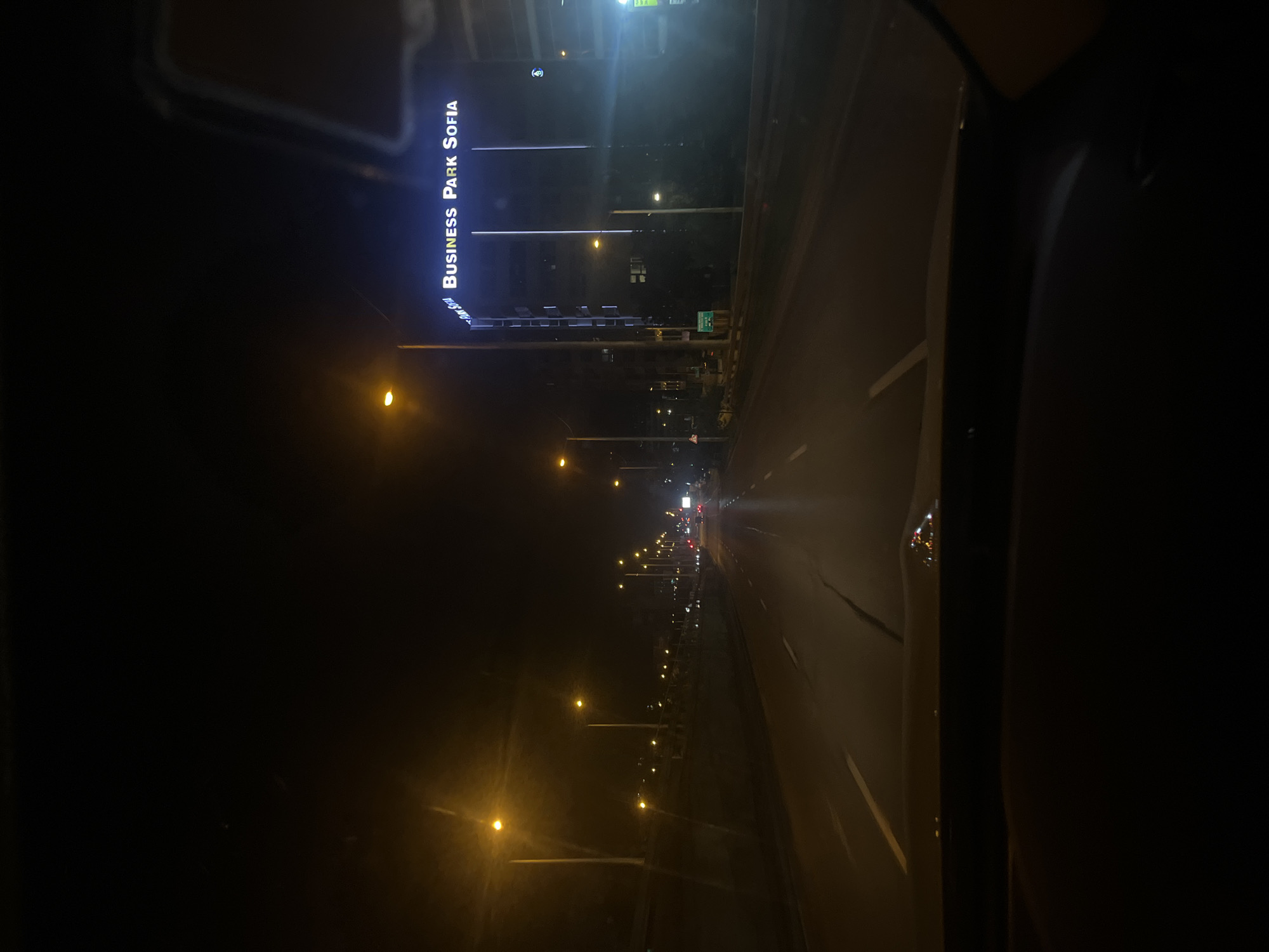 The image depicts a night scene from inside a vehicle traveling on a road in Sofia, Bulgaria. The road is lit by streetlights, and in the distance, there are other vehicles. On the left side of the road, there is a building with a sign that reads "BUSINESS PARK SOFIA." The environment is dark with the primary sources of light being the streetlights and vehicle headlights. There is no visible presence of people in the frame.