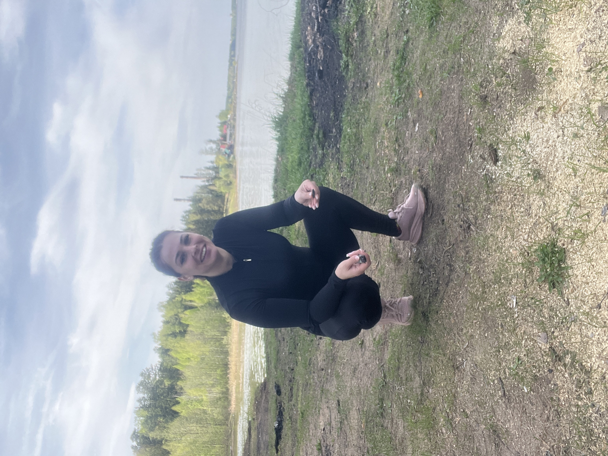 A woman in a black long-sleeved top and leggings is squatting near a body of water, possibly a lake or large pond, in what appears to be a natural setting. She is wearing pink sneakers. The environment includes a mix of grassy and dirt ground cover, with trees and vegetation in the background. The water reflects the sky. The location is the city of Chaikovsky, Russia.