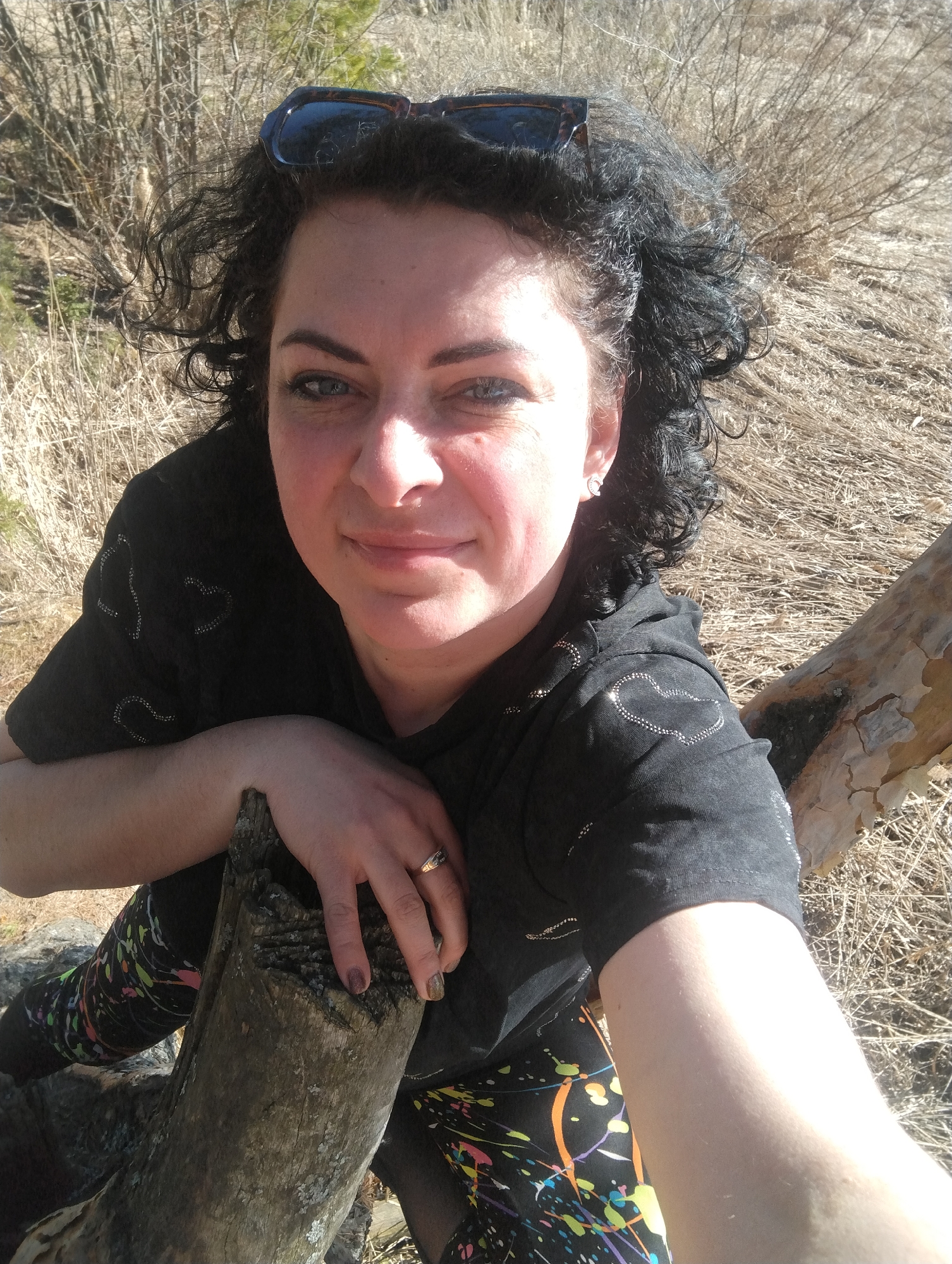 A woman with dark curly hair and sunglasses perched on her head is taking a selfie outdoors. She is leaning on a weathered, light-colored log. She is wearing a black shirt with a heart pattern and colorful leggings. Her left hand, adorned with a gold ring, rests on the log. The background consists of dry grass and bare bushes. There is no indication of Votkinsk or any other city in the image.