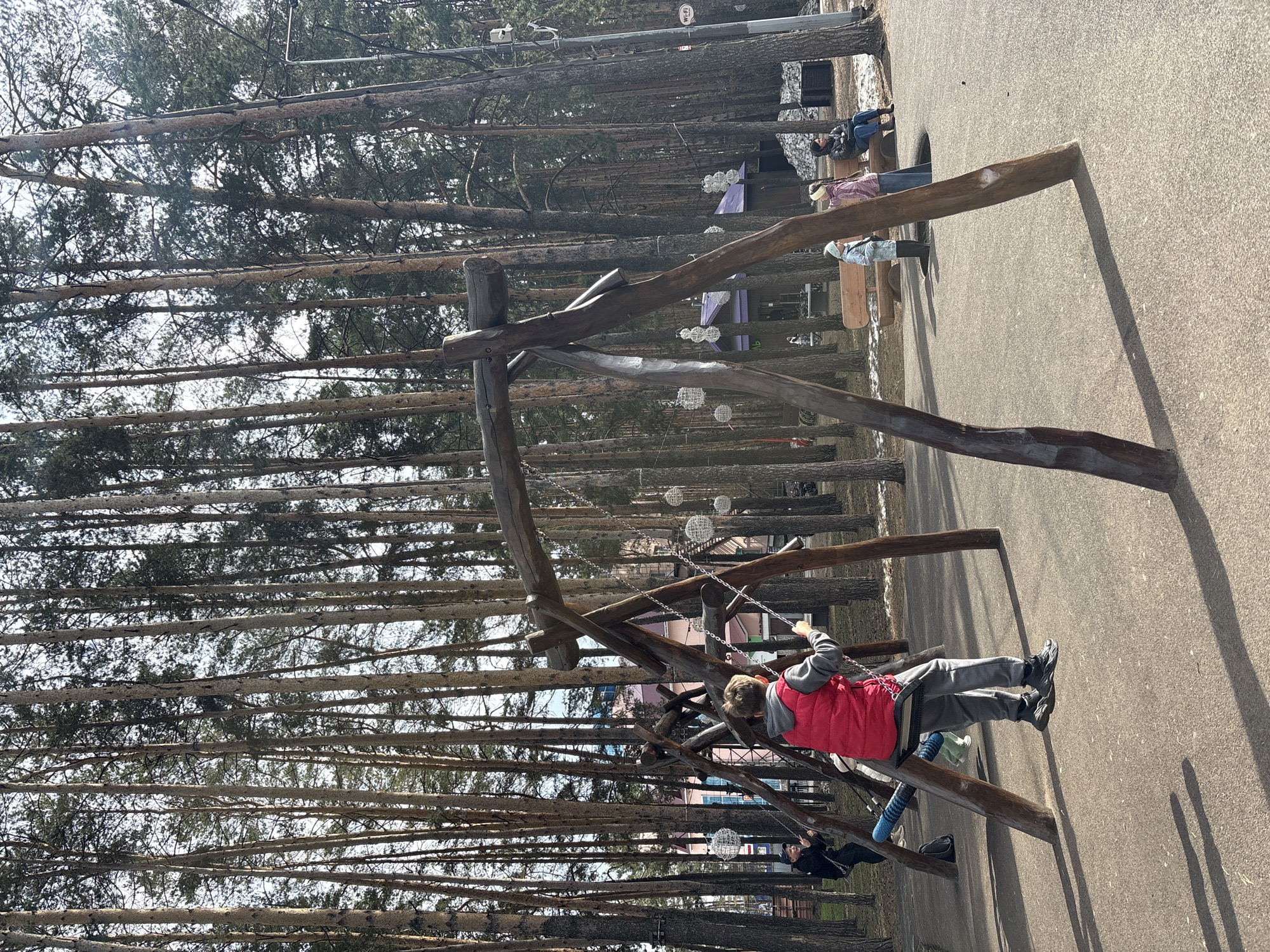 The image shows a playground in Neftekamsk, Russia. The playground features a wooden swing set. A child wearing a red vest and gray pants is swinging on one of the swings. There are other people in the background, sitting on benches. The area is surrounded by tall trees. The ground is paved.