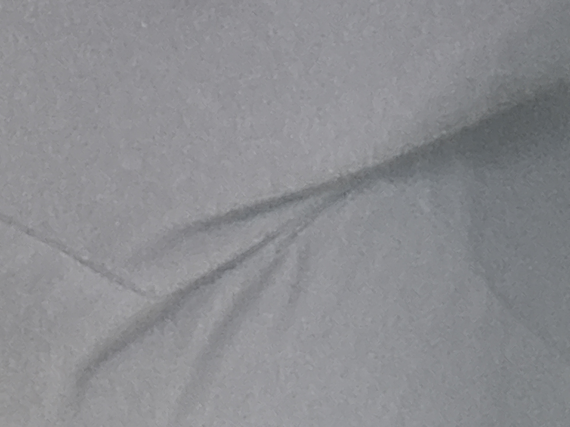 The image shows a close-up, abstract view of a textured grey fabric. The fabric is folded or creased, creating shadows and highlights that give it a three-dimensional appearance. The lighting appears to be soft and diffused. There are no discernible objects or people in the image, nor any indication of a specific location within a city.