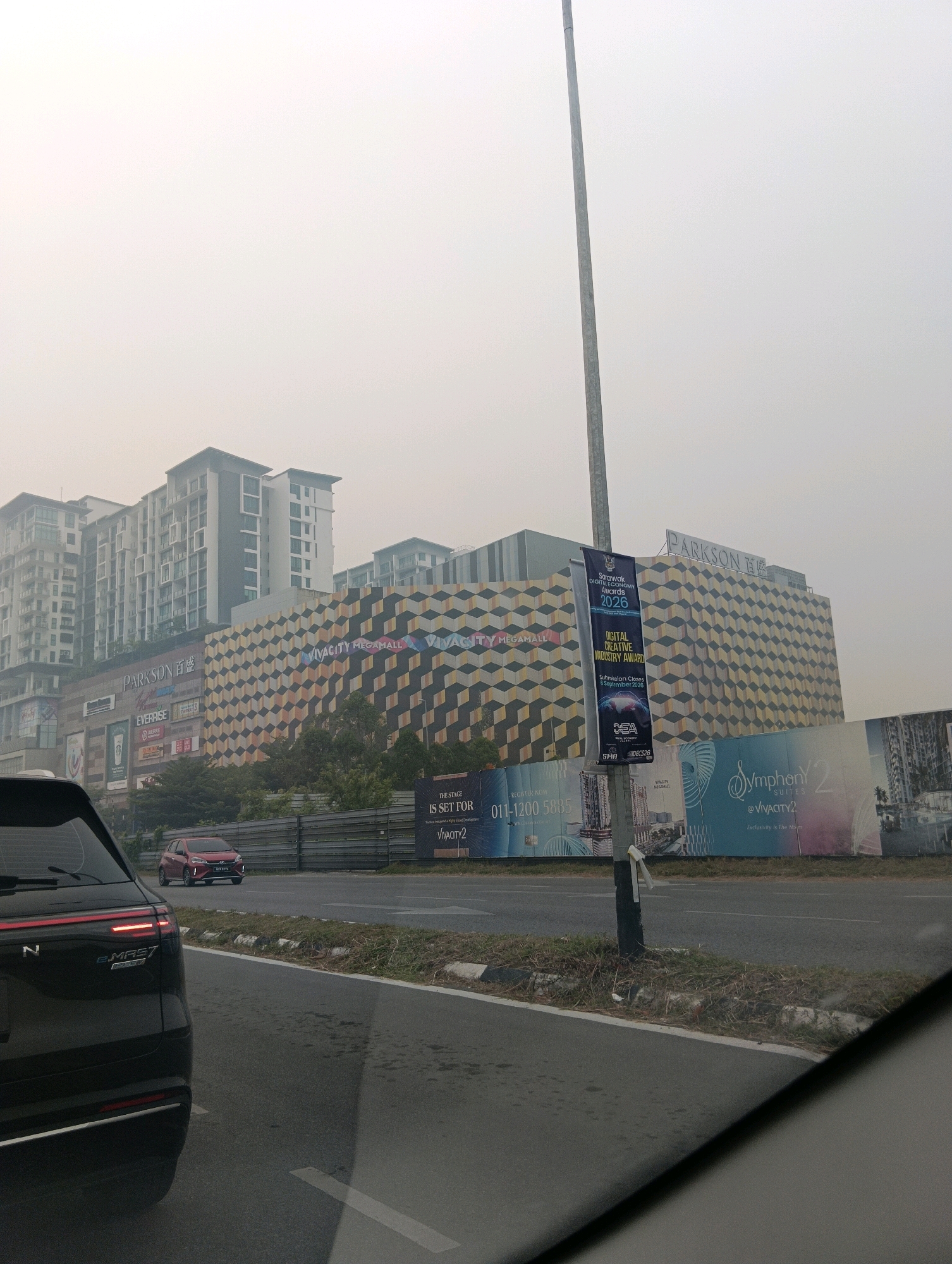 The image depicts a street scene in Kuching, Malaysia, characterized by a hazy or smoky atmosphere. In the foreground, the rear of a dark-colored SUV is visible, suggesting the photo was taken from inside a moving vehicle. On the right side of the road, a red hatchback is observed.
Dominating the background is a large, modern commercial building complex. A prominent sign reads "PARKSON," identifying a department store. Adjacent to it, another building features a distinctive geometric facade with repeating diamond shapes in shades of yellow, orange, and black. Above this facade, the "VIVACITY MEGAMALL" logo is displayed. The architectural style suggests a contemporary urban development.
Several large billboards are positioned along the roadside. One banner prominently displays "Sarawak Digital Economy Awards 2026" and "DIGITAL CREATIVE INDUSTRY AWARD." Another billboard advertises "Symphony Suites @ Vivacity2," with the tagline "Exclusivity Is The Notion." These advertisements indicate a developing commercial and residential area.
The environment appears to be an urban setting, possibly a highway or a major road, with multiple lanes and a grassy median strip. The hazy conditions suggest air pollution or smog. The overall impression is that of a bustling, modern commercial district in Kuching, Malaysia, possibly experiencing adverse atmospheric conditions.