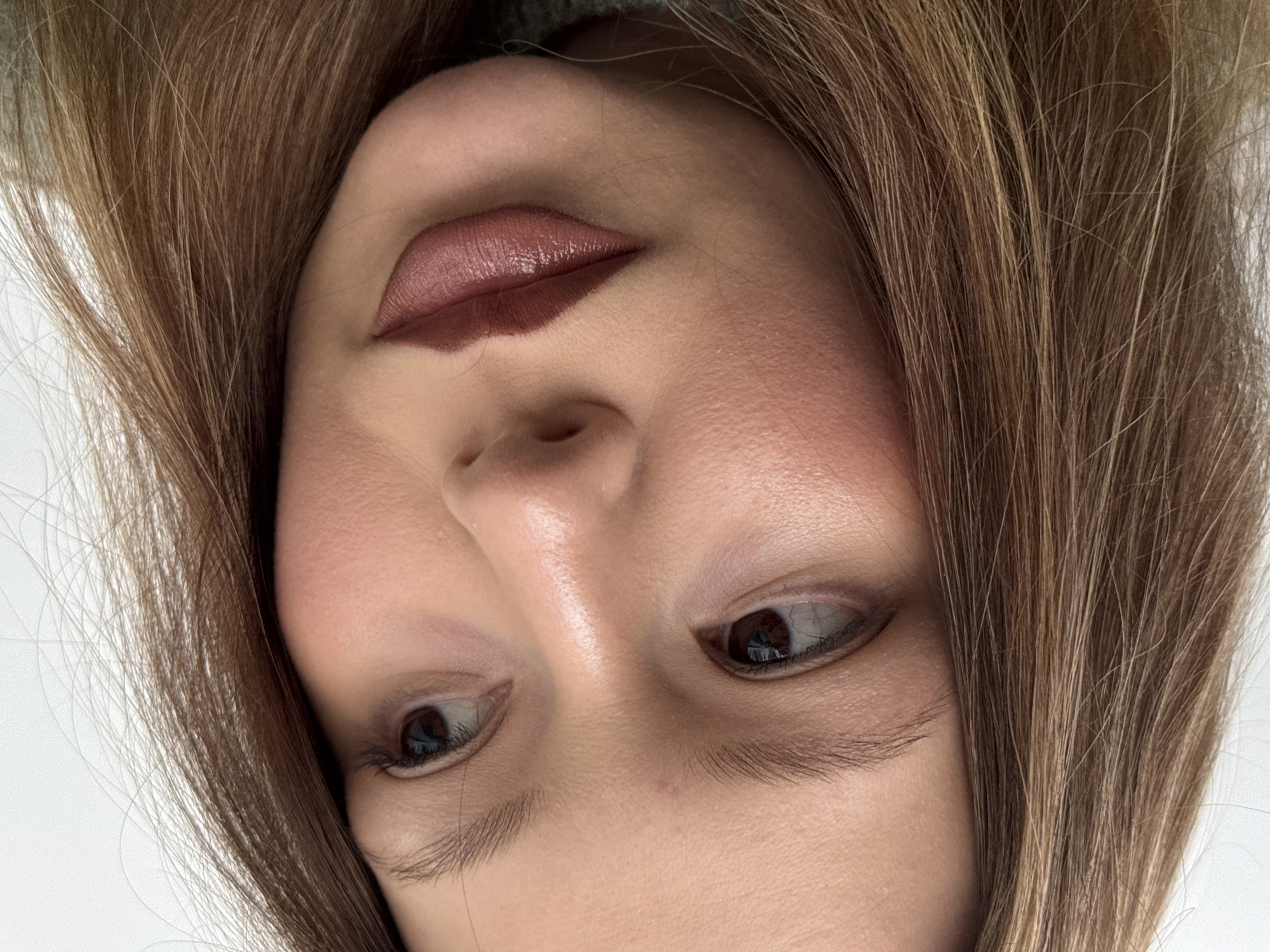 A close-up, upside-down perspective of a woman's face. The woman has light skin, brown hair with lighter highlights, and brown eyes. She is wearing makeup, including lipstick in shades of brown and blush. The background is plain and light-colored. The location is Unknown, Unknown.
