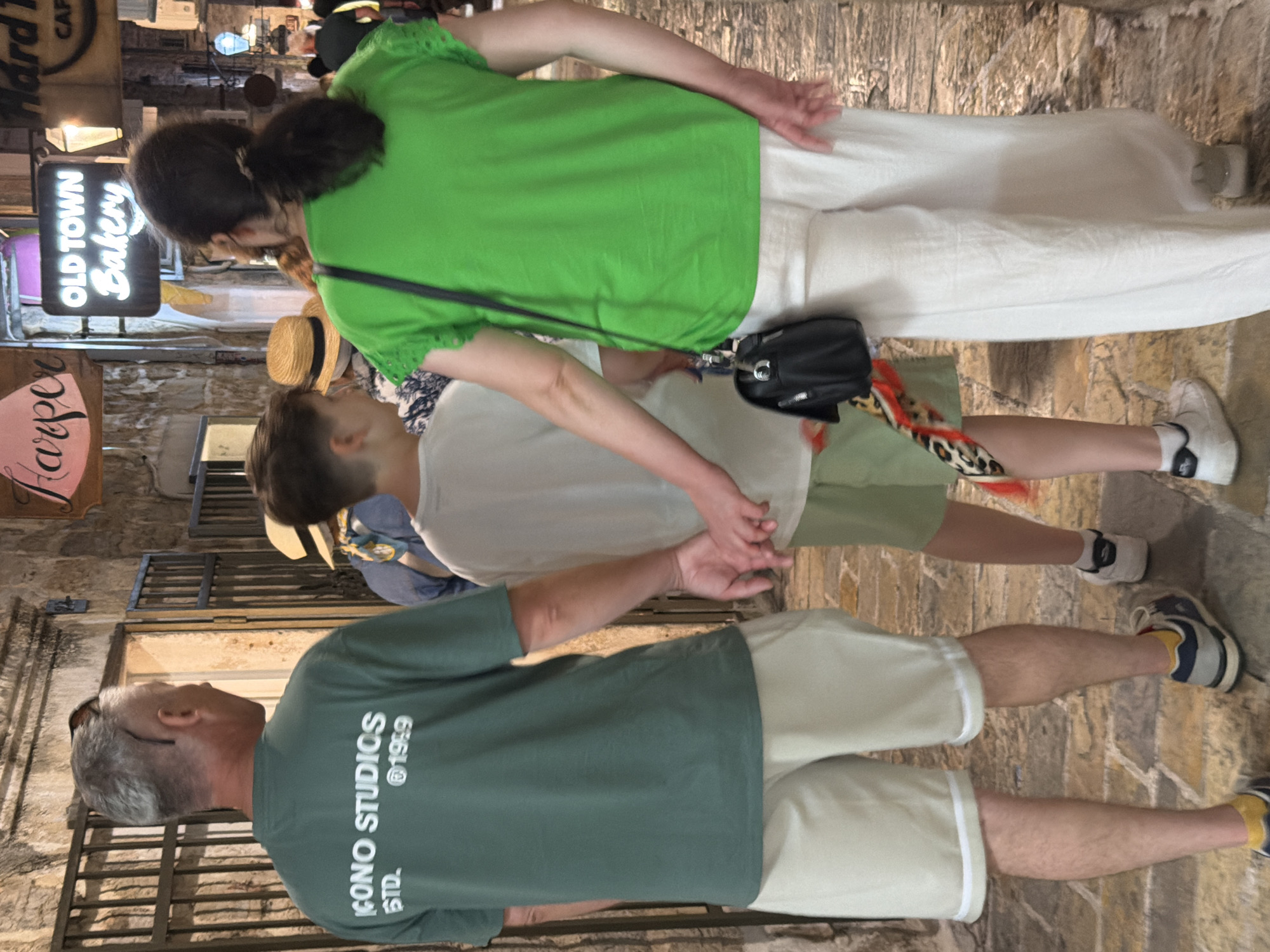 In Budva, Montenegro, a family walks hand-in-hand through a narrow cobblestone alleyway. A man in a green t-shirt and khaki shorts walks on the left, his hand clasped with a boy in a white t-shirt and green shorts in the center. The boy's other hand is held by a woman in a bright green t-shirt and white pants on the right. A black handbag hangs from the woman's shoulder. The background features stone walls, metal gates, and illuminated signs for "Harper" and "Old Town Bakery". The overall atmosphere suggests a casual stroll in a historic urban environment.