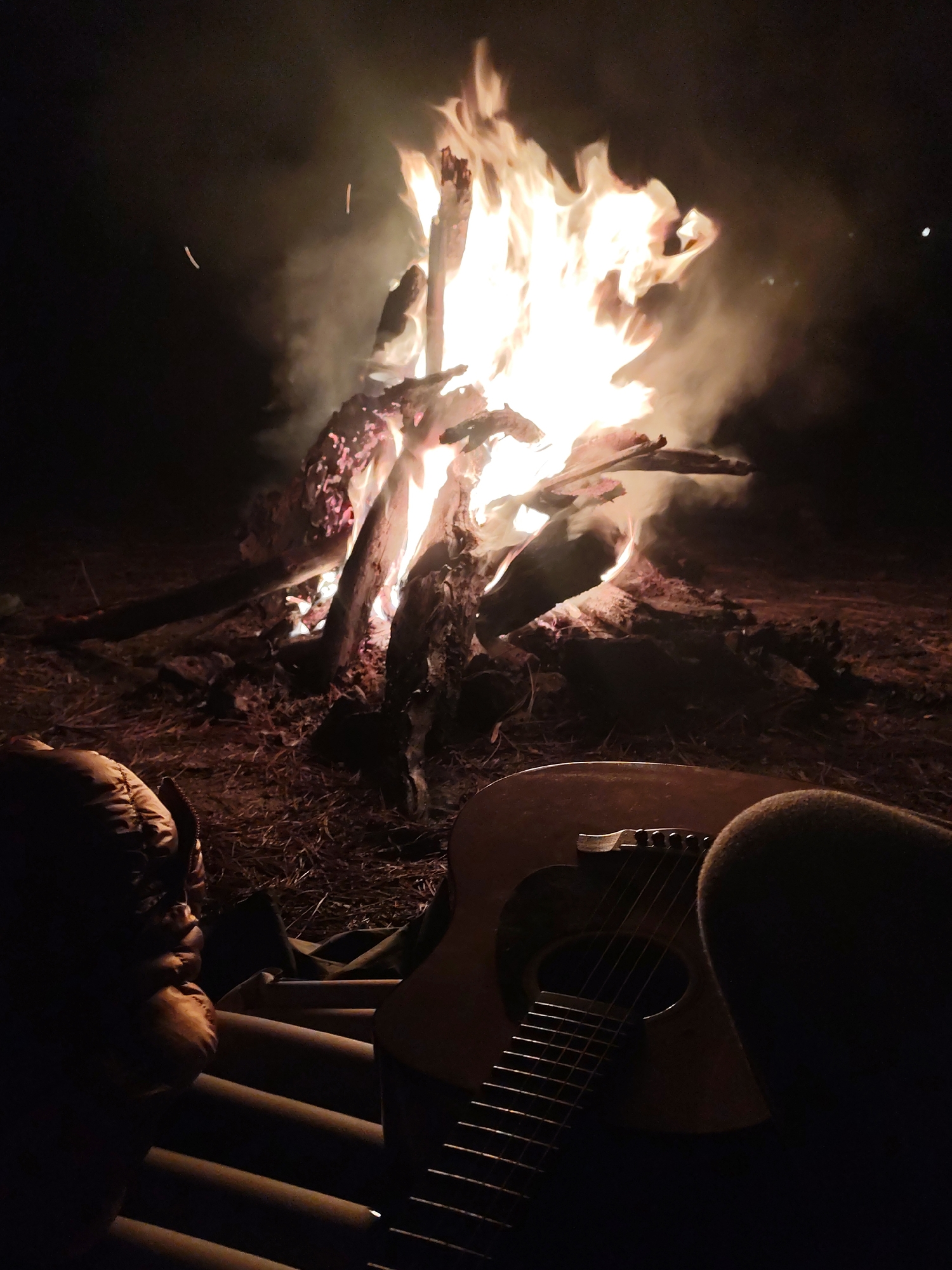 This vertical photograph captures a peaceful nighttime outdoor scene featuring a large, brightly burning bonfire. The roaring flames, a mix of orange and yellow, rise energetically from substantial logs, illuminating the surrounding area with a warm, intense glow. Smoke billows upwards from the fire, creating a hazy backdrop against the dark sky, with a few faint sparks visible near the top left.
In the foreground, an acoustic guitar rests on the ground to the right, its wooden body, soundhole, bridge, and strings clearly visible and gently lit by the firelight. To the left, a person is partially visible, appearing to be seated or reclining on a ribbed mat or sleeping pad, wearing a dark, possibly puffy jacket or sleeping bag. Their position suggests they are facing the fire, enjoying its warmth and ambiance. The ground around the bonfire is covered with natural debris like dry leaves and dirt, indicating a natural, possibly wooded, setting.
The image conveys a sense of tranquility and warmth, characteristic of a campfire gathering. The presence of the guitar suggests that music might be part of the activity, adding to the relaxed atmosphere. The deep darkness of the night sky provides a stark contrast to the vibrant light of the bonfire, highlighting it as the central element and primary light source. There is no visible text in the image.