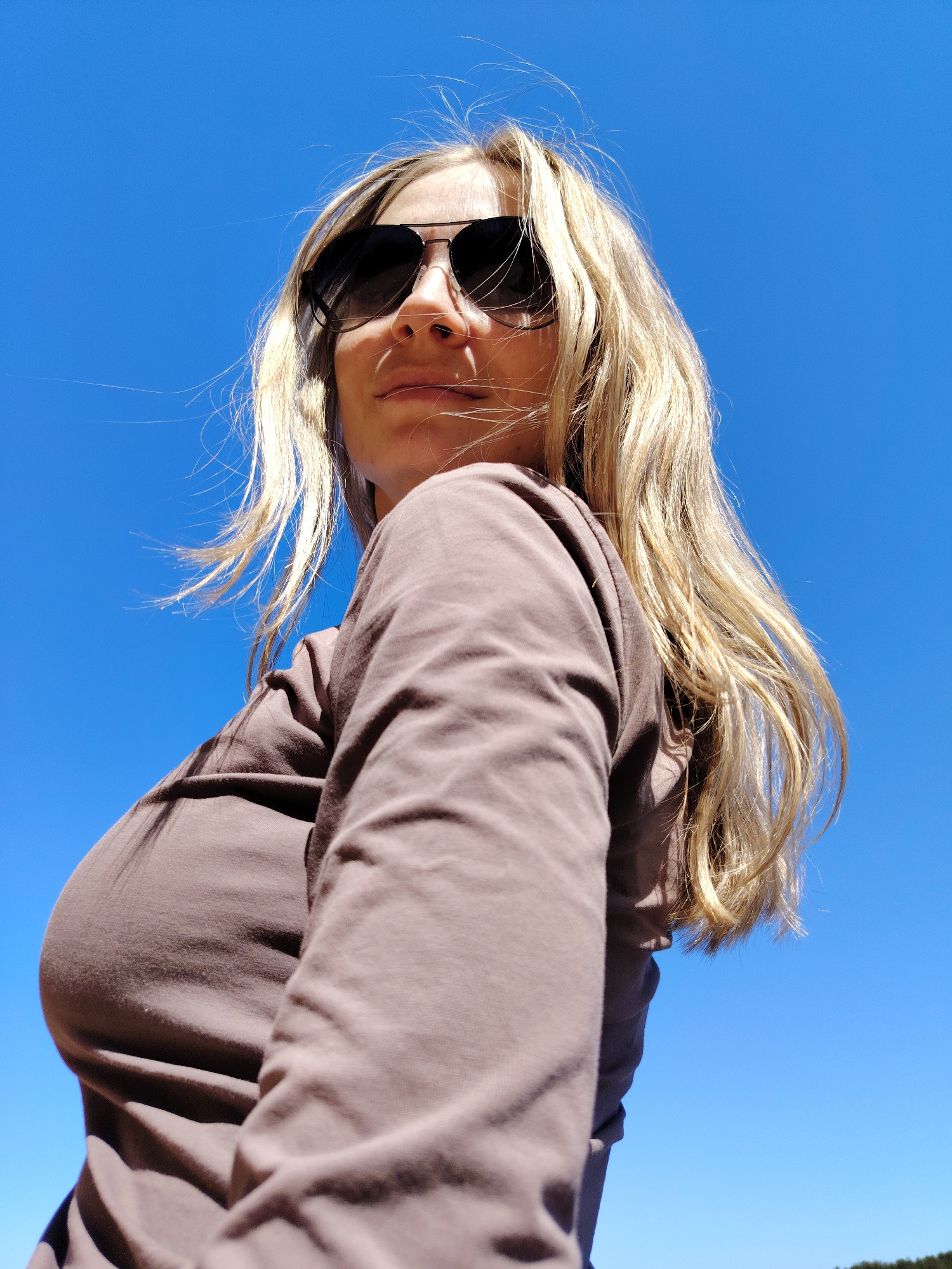 A woman with blonde hair and sunglasses is viewed from a low angle against a bright blue sky. She is wearing a brown long-sleeved shirt. The location in Chaikovsky, Russia, is not evident from the image.