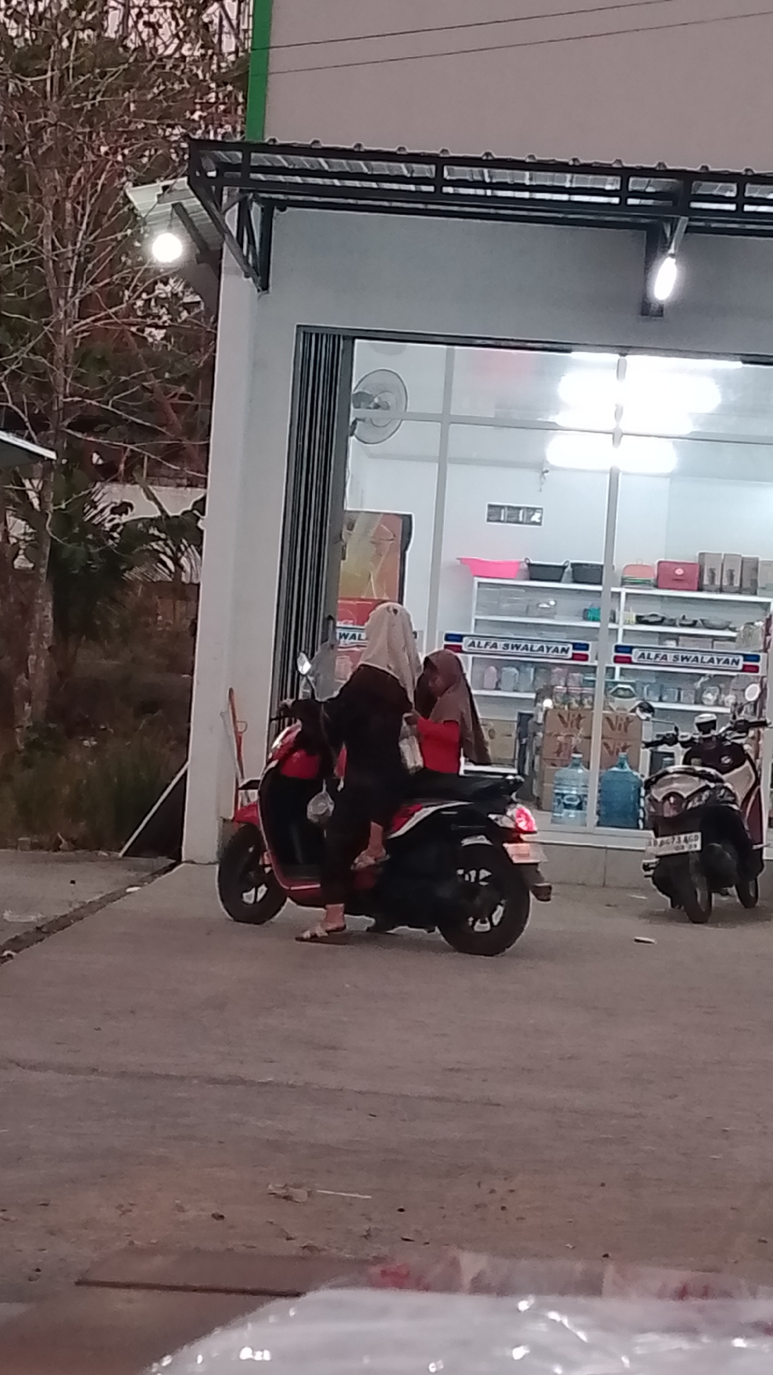 In Karanglangu, Indonesia, two individuals are positioned on a red scooter parked in front of a store. The person in the driver's seat wears a black abaya and a white hijab, while the passenger in the back wears a red shirt and a brown hijab. The scooter's kickstand is down, and it is parked on a paved surface. Behind the scooter, the store has large glass windows displaying shelves stocked with various goods, including water bottles and boxed items. The store's signage reads "ALFA SWALAYAN". Another scooter, predominantly black, is parked to the right of the red scooter, also on the paved surface. The environment suggests an evening or dusk setting, with artificial lighting illuminating the area in front of the store. Bare tree branches are visible to the left of the frame.