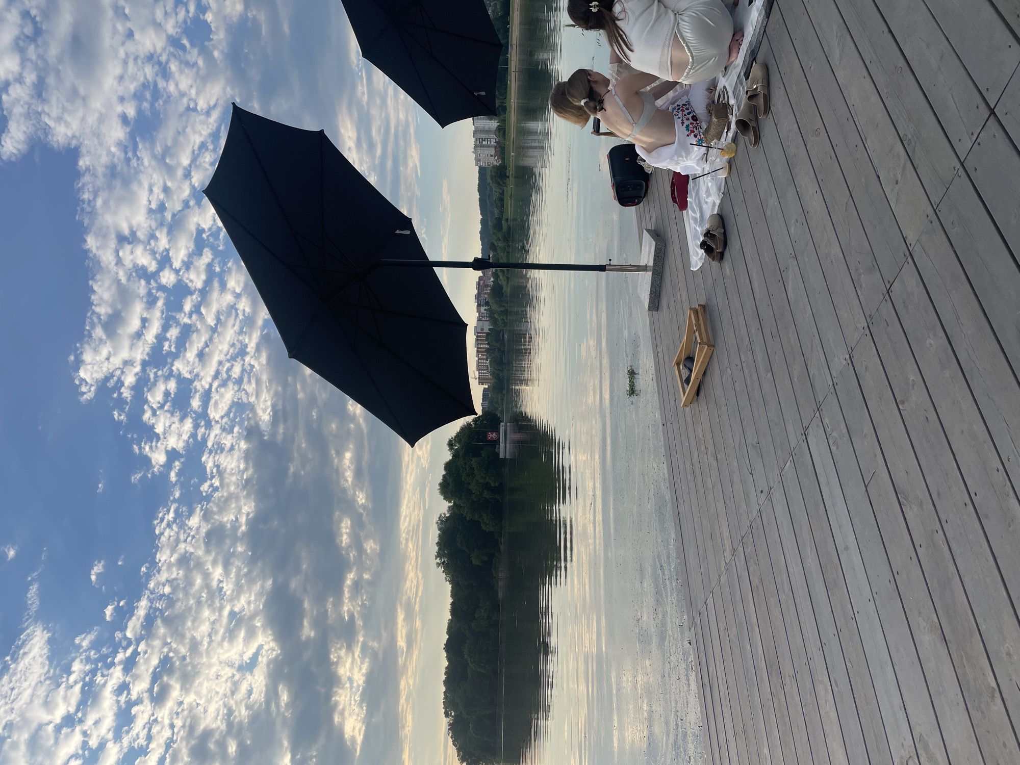 On a wooden pier beside a body of water, two individuals are seated, facing away from the camera. They appear to be engaged in a casual gathering or picnic, with a blanket spread out on the pier and a drink visible on the blanket. Two large black umbrellas are positioned to provide shade. In the background, a tree-lined shore is visible across the water, with buildings and a cloudy sky above. The scene suggests a relaxed outdoor setting, possibly during the late afternoon or early evening. The specific location of Khmelnytskyi, Ukraine, is not visually discernible from the image itself, as there are no unique landmarks or identifying features that definitively place it in that city.