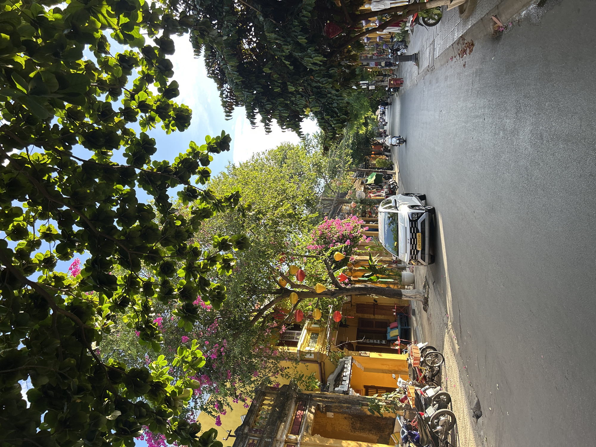 The image displays a daytime street scene in Hội An Ward, Vietnam. Overhead, a dense canopy of large green tree leaves is present, with patches of bright blue sky and some white clouds visible through the foliage. Sunlight filters through the leaves, creating dappled lighting.
On the left side of the foreground, several motorbikes are parked on a concrete sidewalk, which has an uneven surface and a visible drain or pothole. Adjacent to the motorbikes, a white Hyundai SUV is parked, facing away from the camera, with its front right wheel partially on the sidewalk. The vehicle's license plate reads "32H-053.60".
To the left of the street, a row of multi-story buildings, predominantly painted vibrant yellow, lines the road. These buildings feature traditional architecture with tiled roofs, ornate balconies, and wooden shutters. One prominent tree in front of a yellow building is adorned with both pink bougainvillea flowers and numerous red and yellow traditional Vietnamese lanterns. Some buildings have small outdoor seating areas with tables and colorful cushions.
The asphalt road surface is visible, showing some debris and fallen flowers near the curb. A single individual on a white scooter, wearing a white helmet, is driving down the center of the street, moving away from the viewer. Further down the street, more pedestrians and motorbikes are visible.
On the right side of the street, another line of buildings, some displaying merchandise like mannequins, is present, interspersed with tall, dark green trees forming a denser canopy than on the left. A black lamppost and a red fire hydrant are visible on the right sidewalk. The street recedes into the distance, lined by trees and buildings, suggesting a continued urban environment.