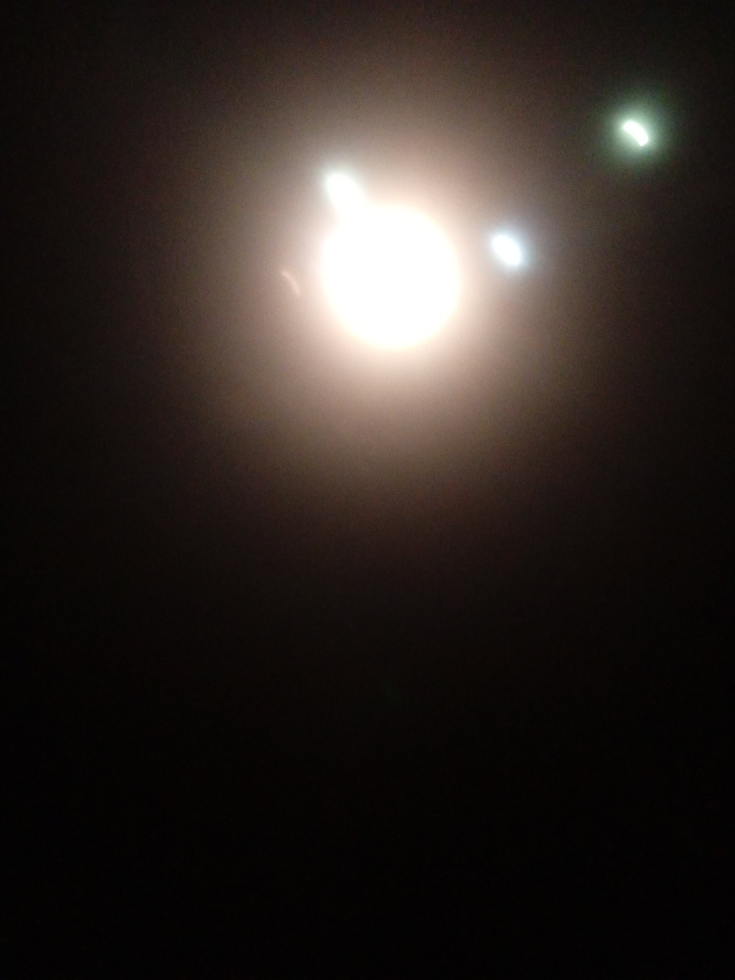 The image is a dark, out-of-focus photograph with three bright, circular light sources visible against a black background. The largest light source is in the upper left quadrant, appearing white and slightly diffused. To its right and slightly lower, there is a smaller, white light source. Further to the right and slightly higher than the second light source, there is a third, greenish-white light source, also circular. The overall impression is of a nighttime scene, possibly with some atmospheric haze or dust. There are no discernible people, objects, or environmental details beyond the light sources and the dark background. The location in Sabon Gari Nassarawa, Nigeria, is not visually evident from the image itself.