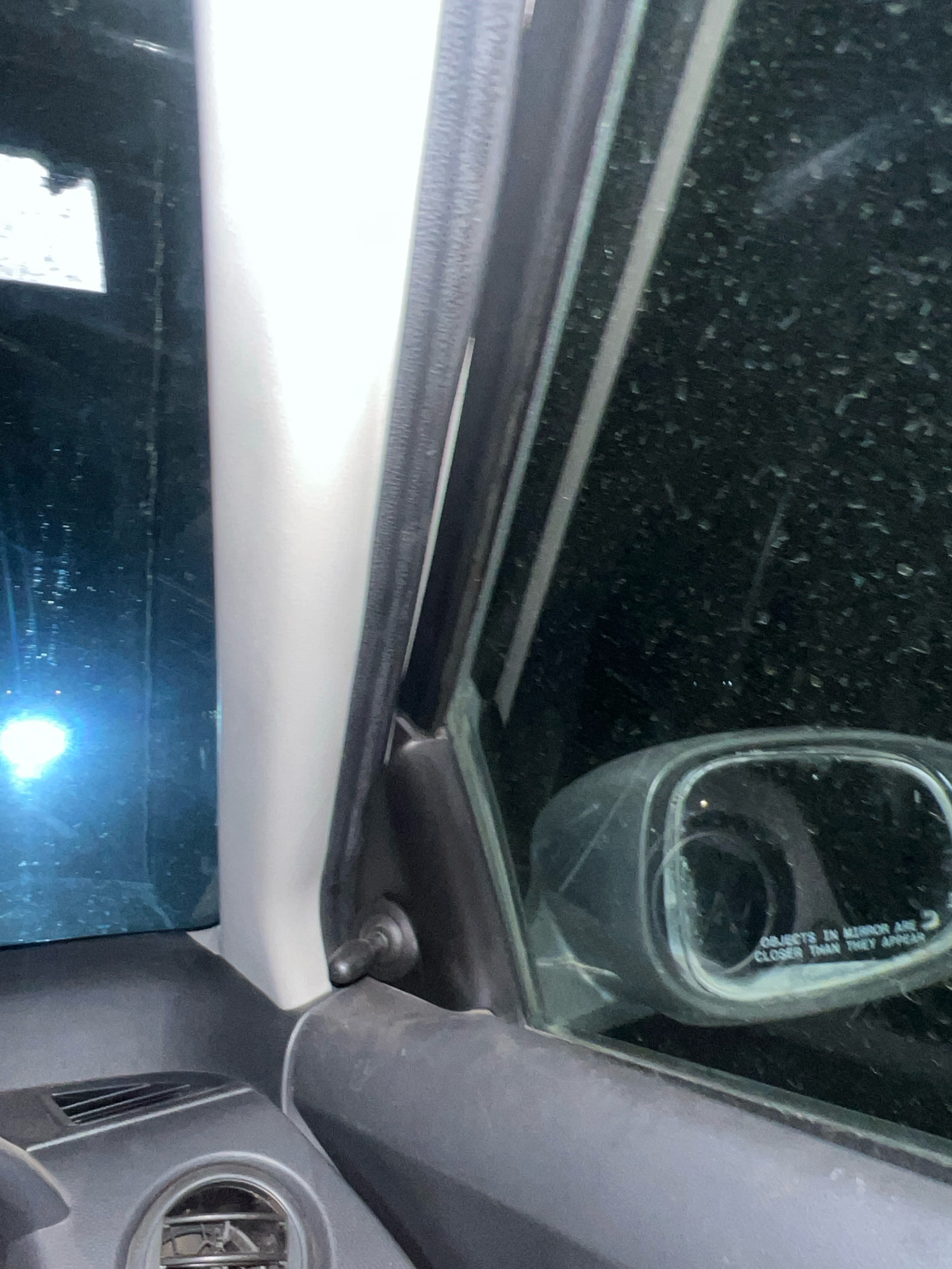 This image captures a close-up view of the interior of a vehicle, likely a car. The perspective is from the driver's seat, looking towards the side window and side-view mirror.
The dominant elements are the car's interior trim, the window, and the side-view mirror. The trim appears to be made of dark plastic, with a lighter, possibly painted, A-pillar separating the windshield and side window. The side window is tinted and reflects some light, showing a blurry, out-of-focus light source on the left. The side-view mirror is visible, with the standard "objects in mirror are closer than they appear" text inscribed on it. The exterior seen in the mirror is dark and appears to have a textured surface, possibly with raindrops or dirt.
The image also shows parts of the car's dashboard, including an air vent and a circular vent or control knob. The lighting suggests it might be nighttime or a dimly lit environment, with the interior lights of the car potentially being on.
The scene is located within the city of Kum, India. This specific interior shot does not contain any explicit landmarks or environmental cues that can pinpoint a more precise location within Kum. The focus is on the immediate interior of the vehicle. There are no people visible in the image. The objects are stationary.