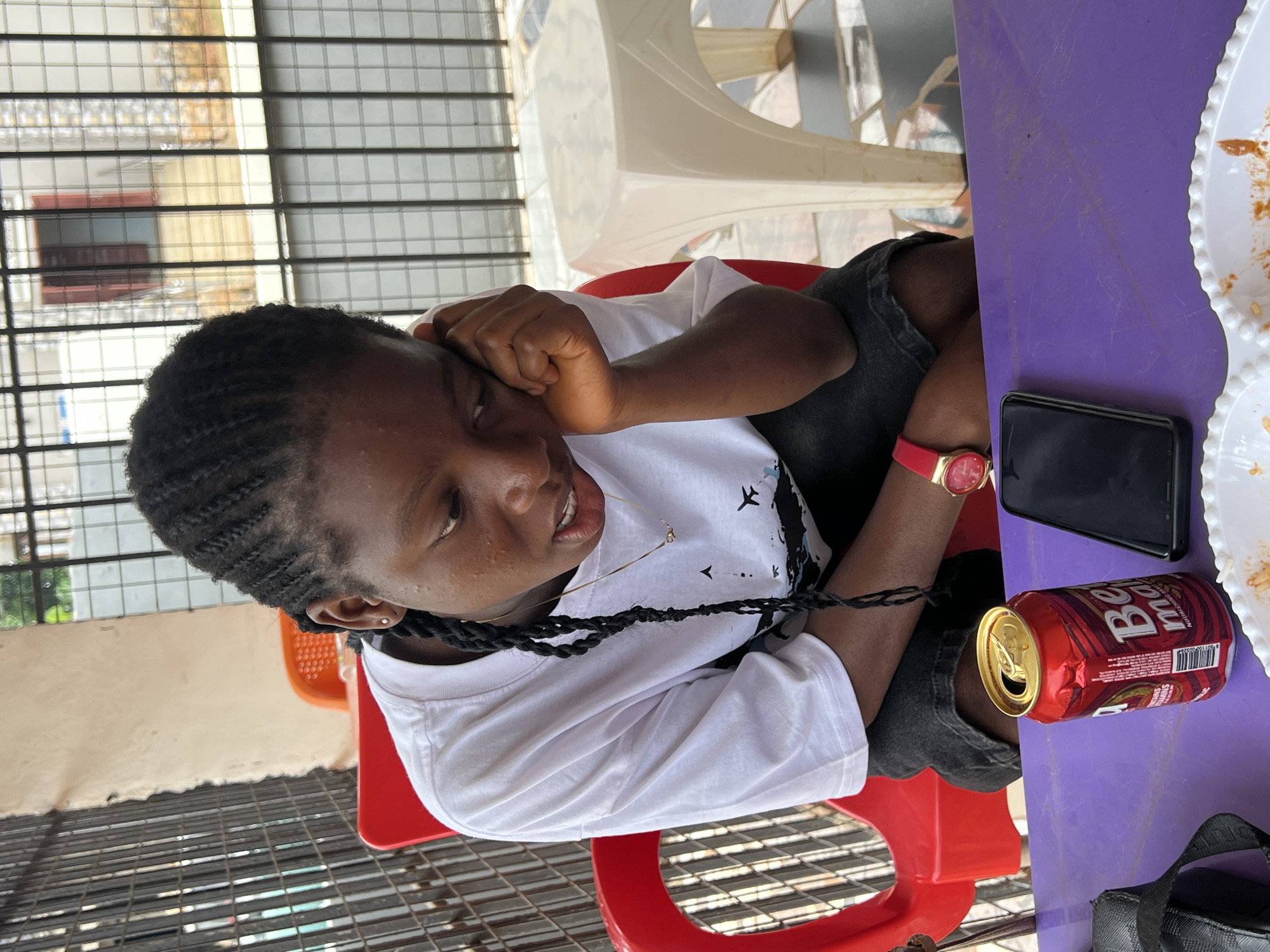 A young Black person with braided hair rests their head on their right hand, looking down with a pensive expression. They are wearing a white t-shirt with a graphic print and dark shorts, and a red wristwatch on their left wrist. In front of them on a purple table are an open red soda can and a black smartphone. The scene appears to be outdoors, possibly at a roadside eatery or bar in Nkpor, Nigeria, with a metal fence and a building visible in the background. A red plastic chair is to the left of the person, and a white plastic stool is to the right.