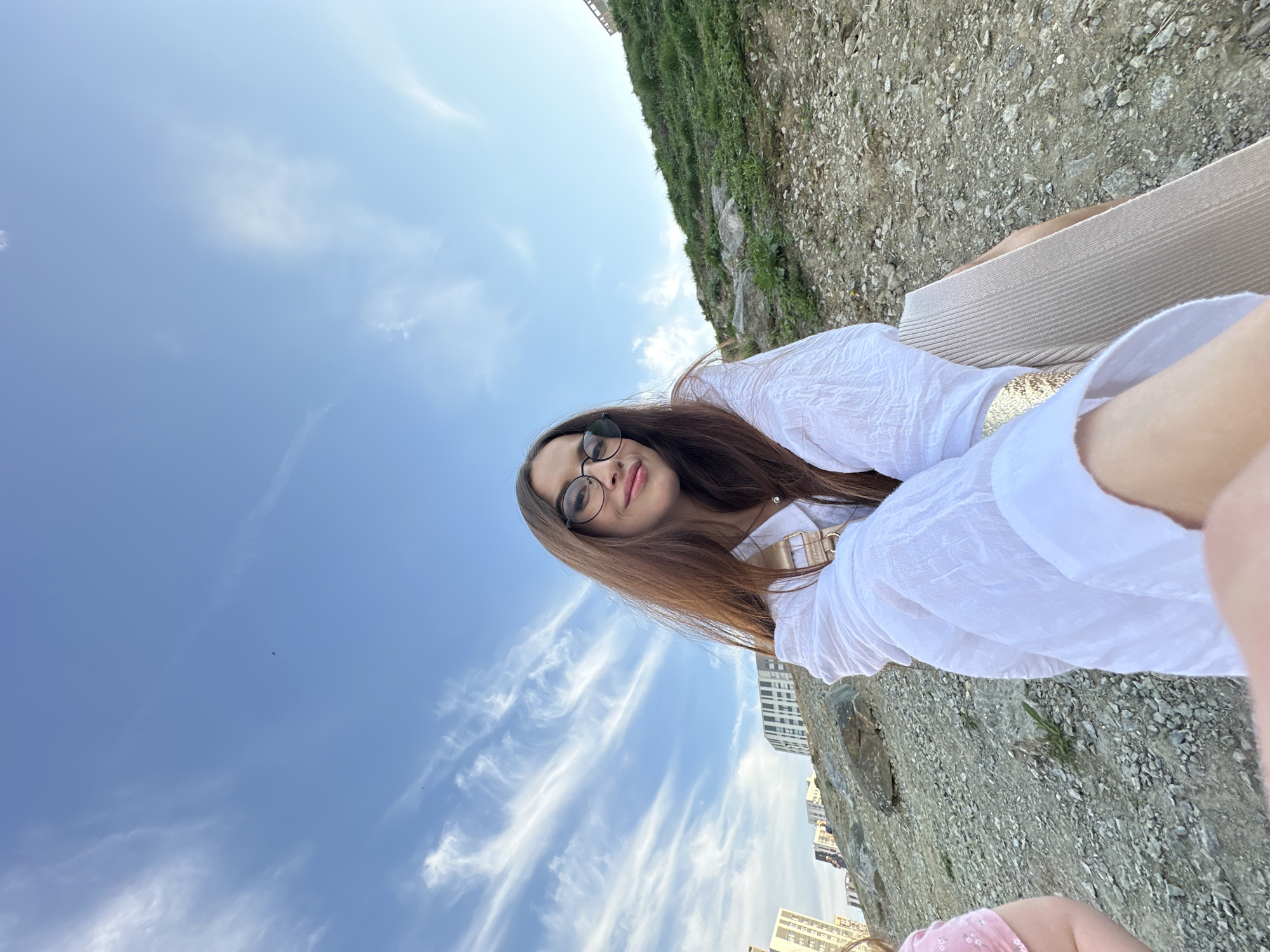 A rotated medium shot captures a woman with long brown hair and glasses, wearing a white shirt with a gold belt. She is positioned in front of a blue sky with wispy clouds. Behind her, there is a steep embankment covered in vegetation and rough terrain. In the background, there are several tall buildings, indicative of an urban environment. The photo appears to be taken in Yekaterinburg, Russia.