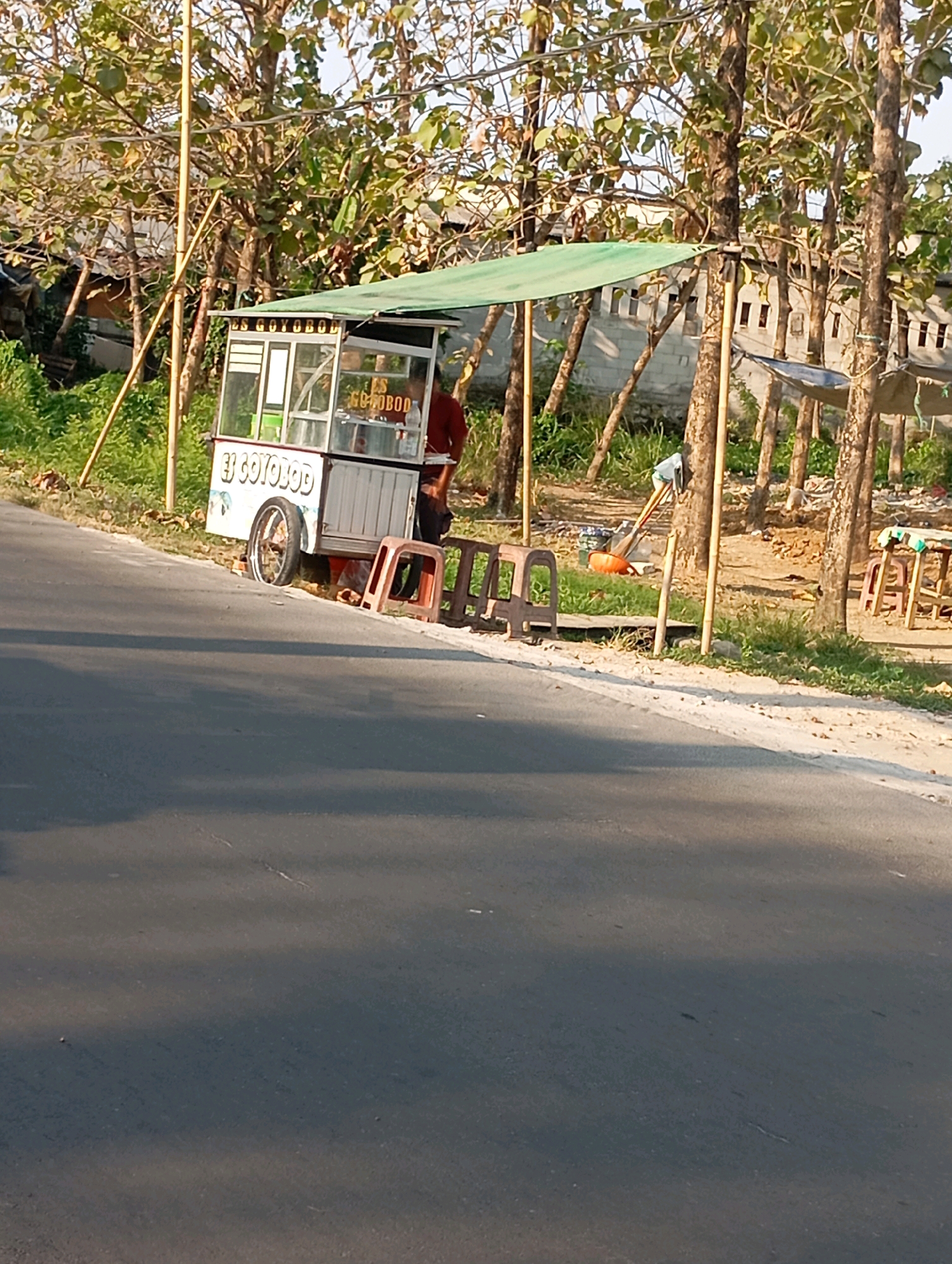 The image shows a street scene in Babelan, Indonesia, dominated by a food cart selling "ES GOTOBOB" in front of a line of trees. The food cart is white with green trim and has a canopy. A person in a maroon shirt is standing behind the cart. Two red plastic chairs are placed on the side of the road near the cart, and a broom and dustpan are leaning against a tree. The road in the foreground is paved and appears to be a two-lane street. In the background, there are buildings with windows and a fence. The scene is bathed in daylight, casting shadows on the road.